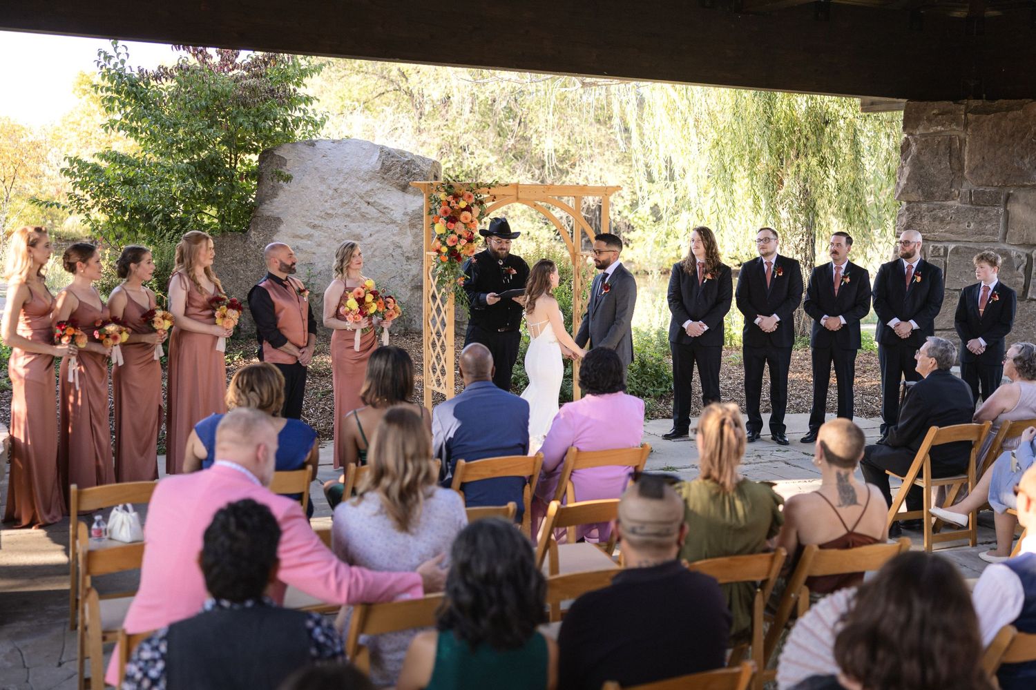 Outdoor wedding ceremony under a stone pavilion with guests seated on folding chairs.
