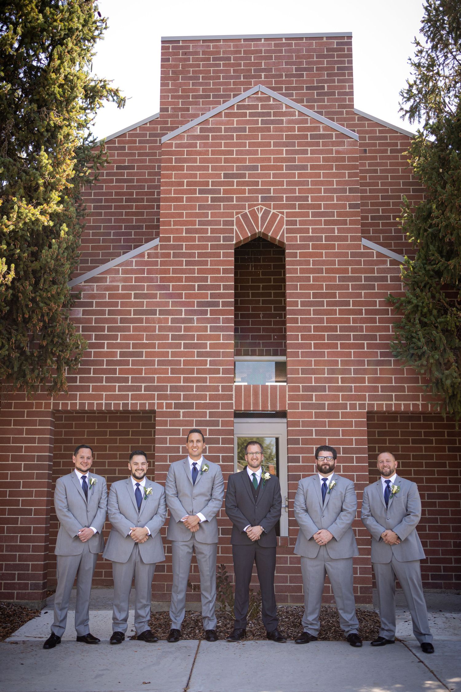 Groomsmen in matching gray suits pose in front of a brick church building.