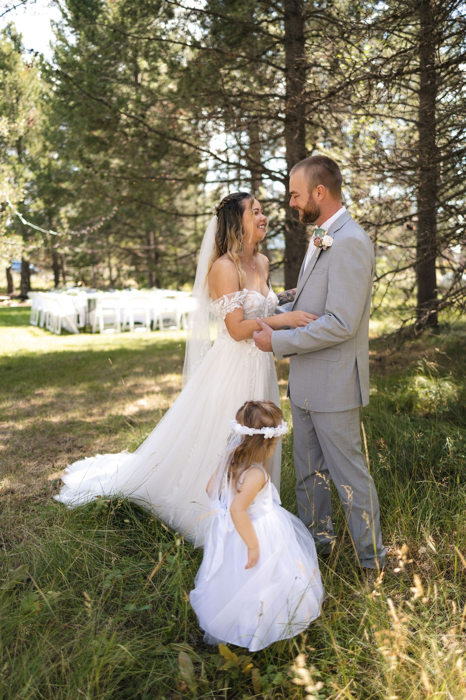 A couple shares an intimate moment in their wedding attire while a small flower girl joins them in a forest setting.