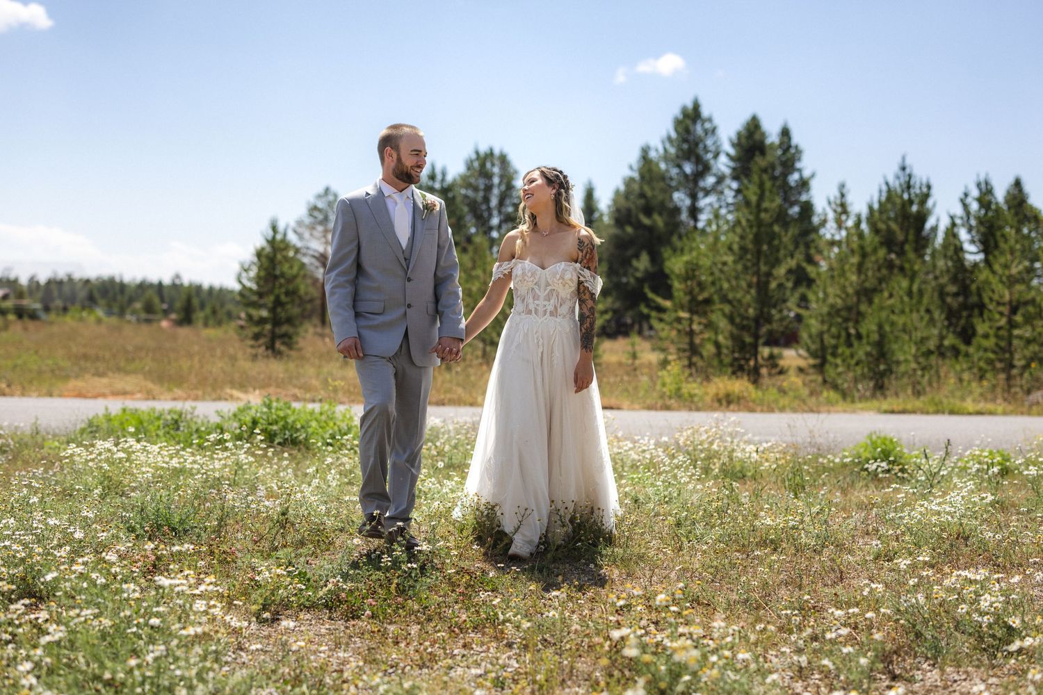 Bride and groom stand together in casual pose amidst wildflower field with pine forest backdrop.