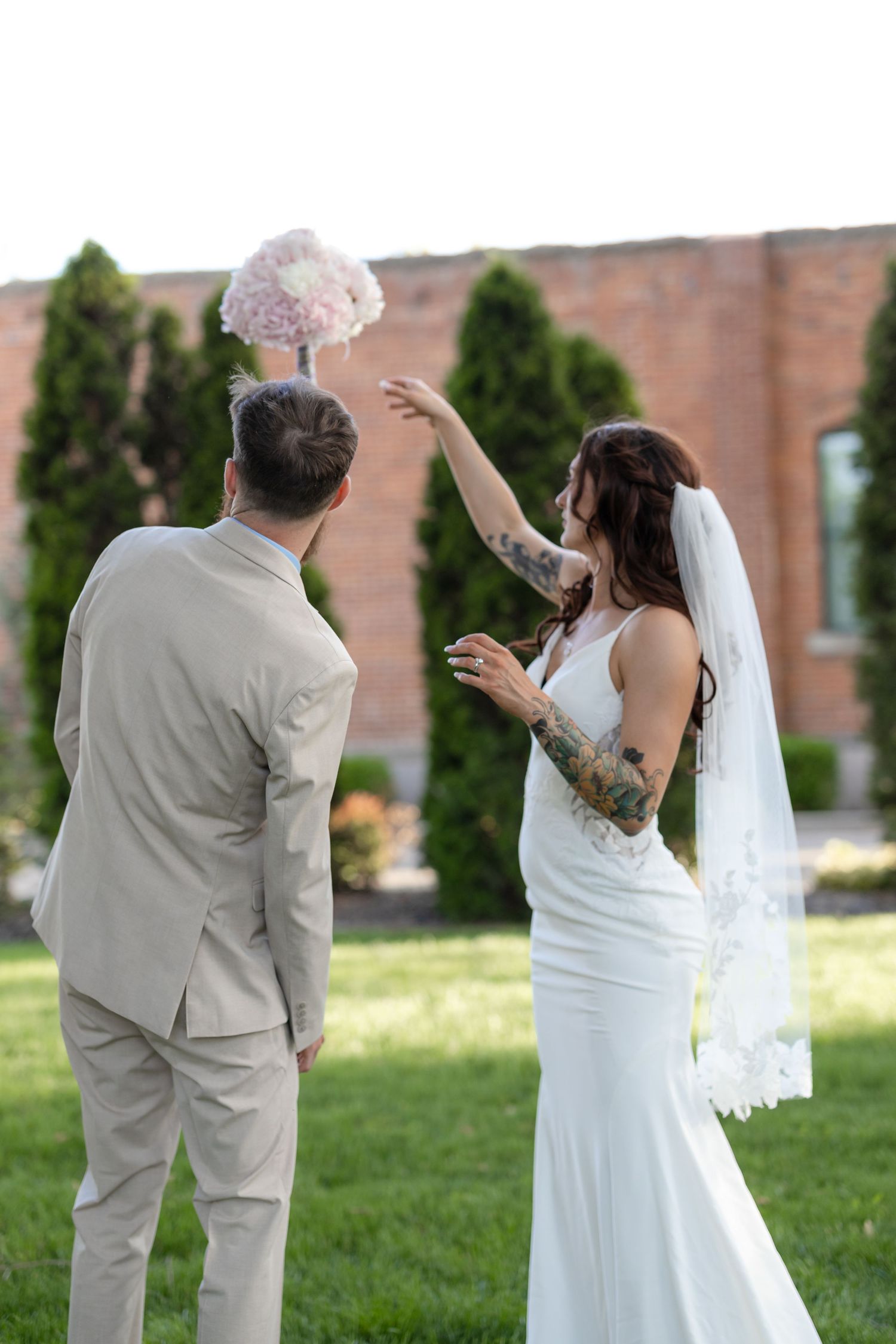 Wedding couple on grass with brick building background playfully toss pink bouquet in the air.
