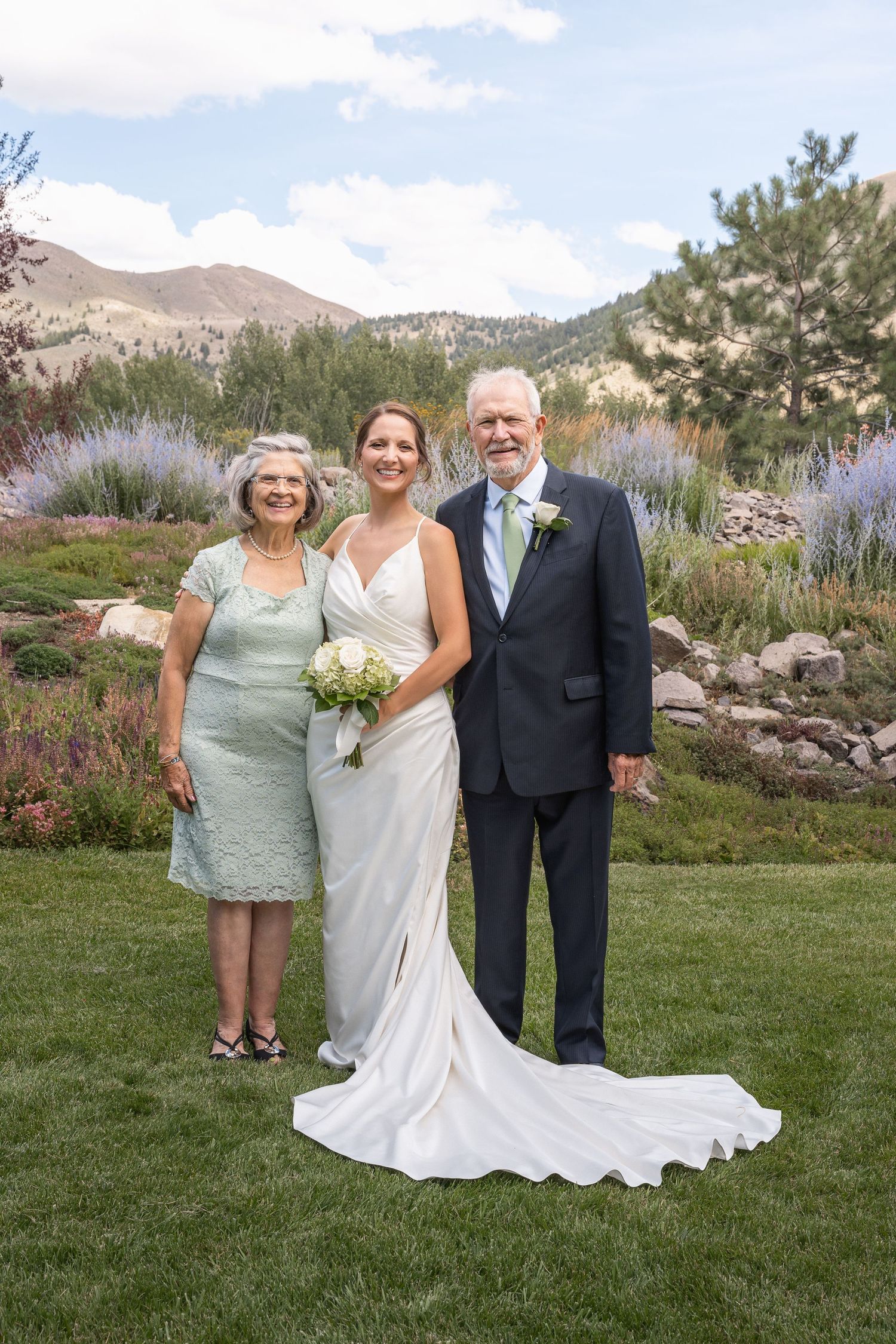 Wedding group poses on manicured lawn with mountain scenery and purple flowers in background.