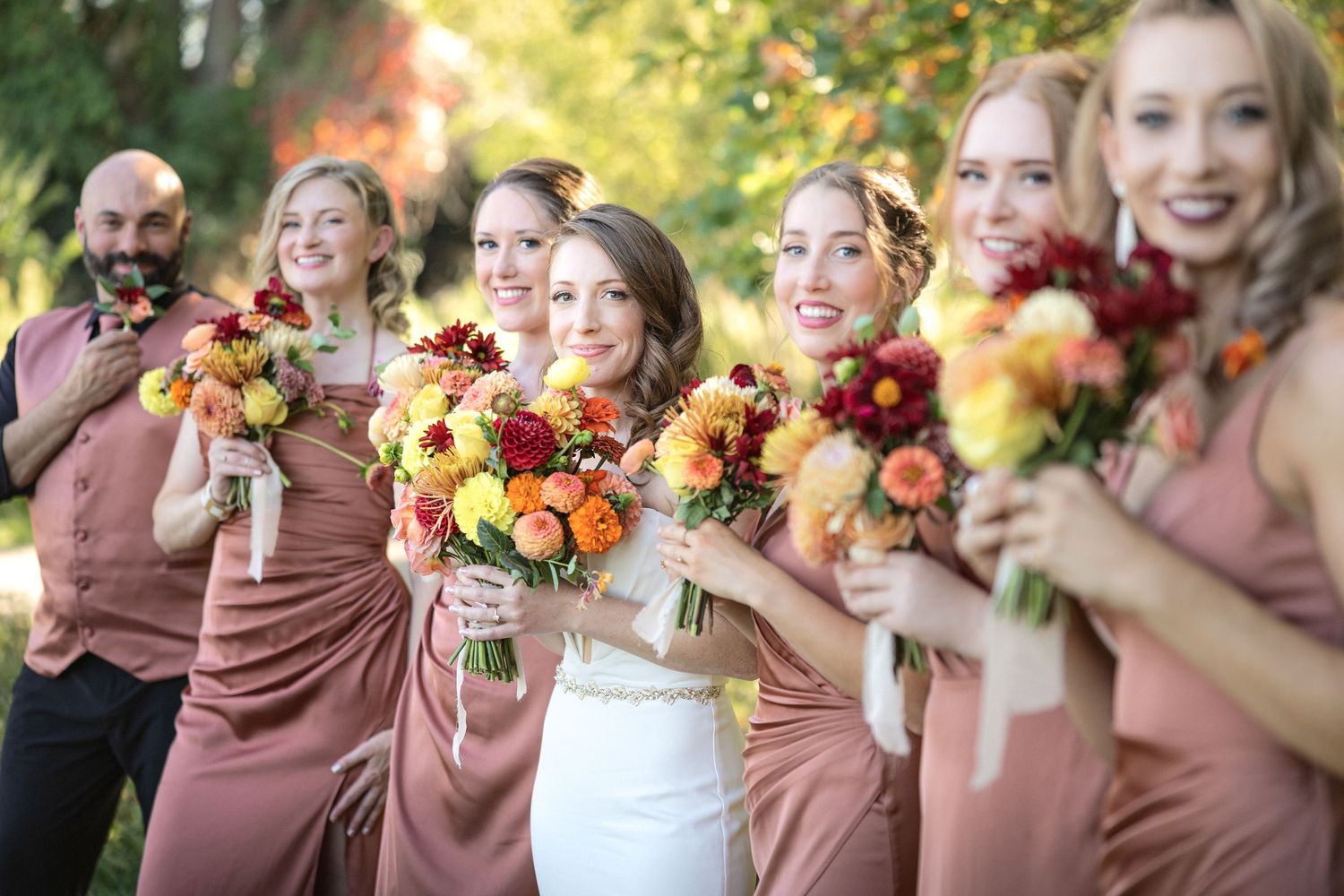 Bridesmaids in dusty rose dresses hold vibrant autumn bouquets.