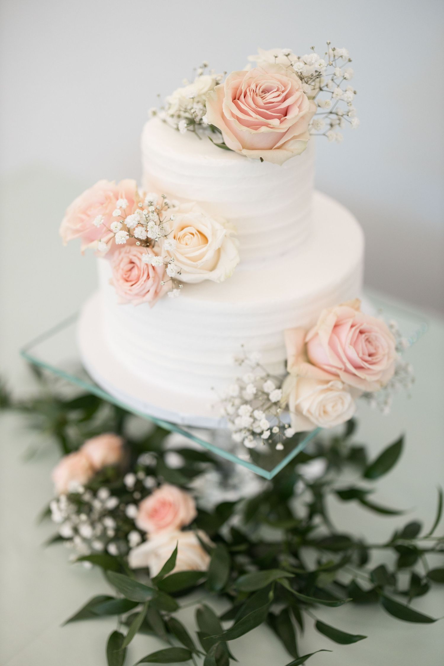 White wedding cake decorated with delicate pink roses and baby's breath on a marble surface with greenery accents.