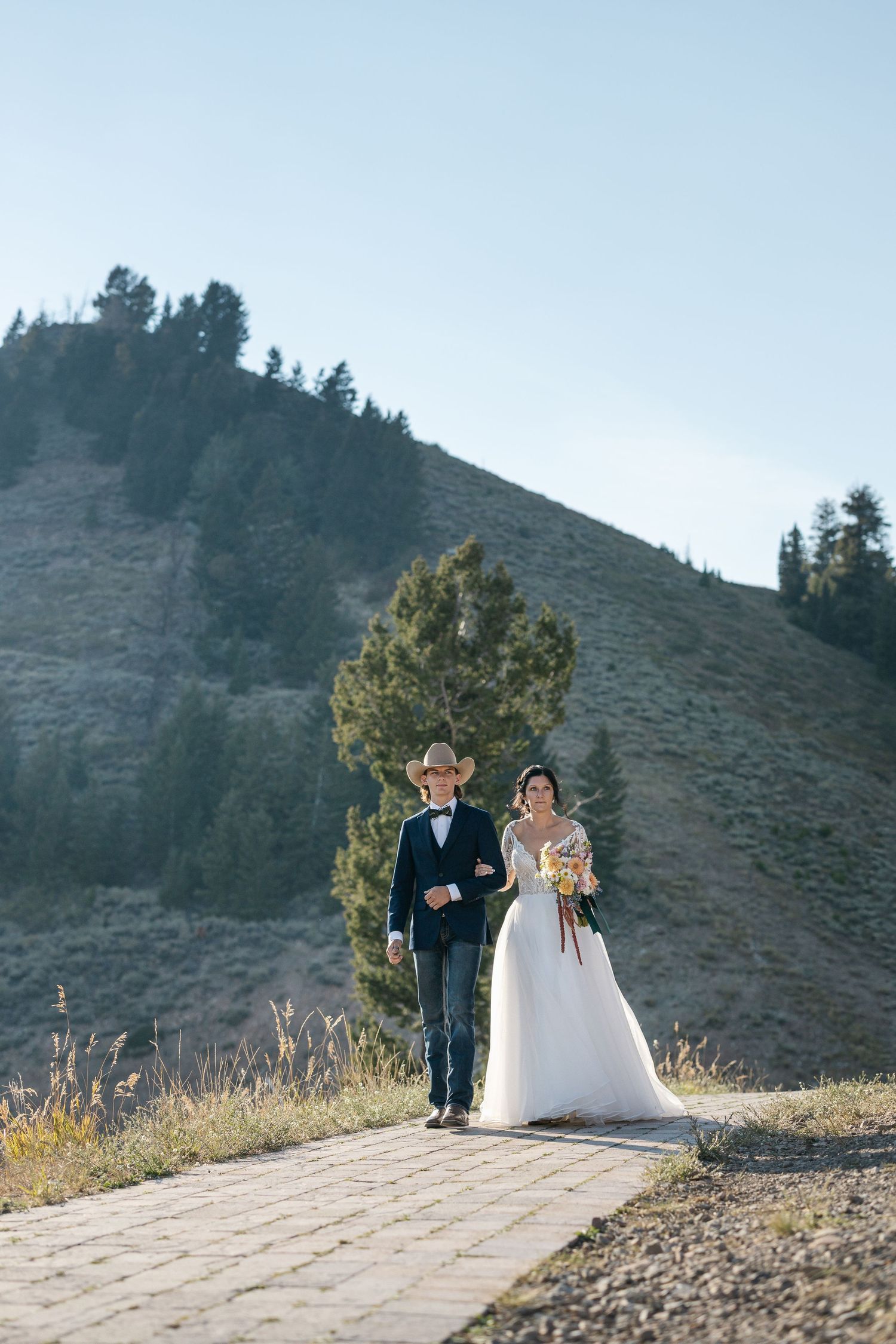 Wedding couple stands on stone path against backdrop of mountain slopes and blue sky.