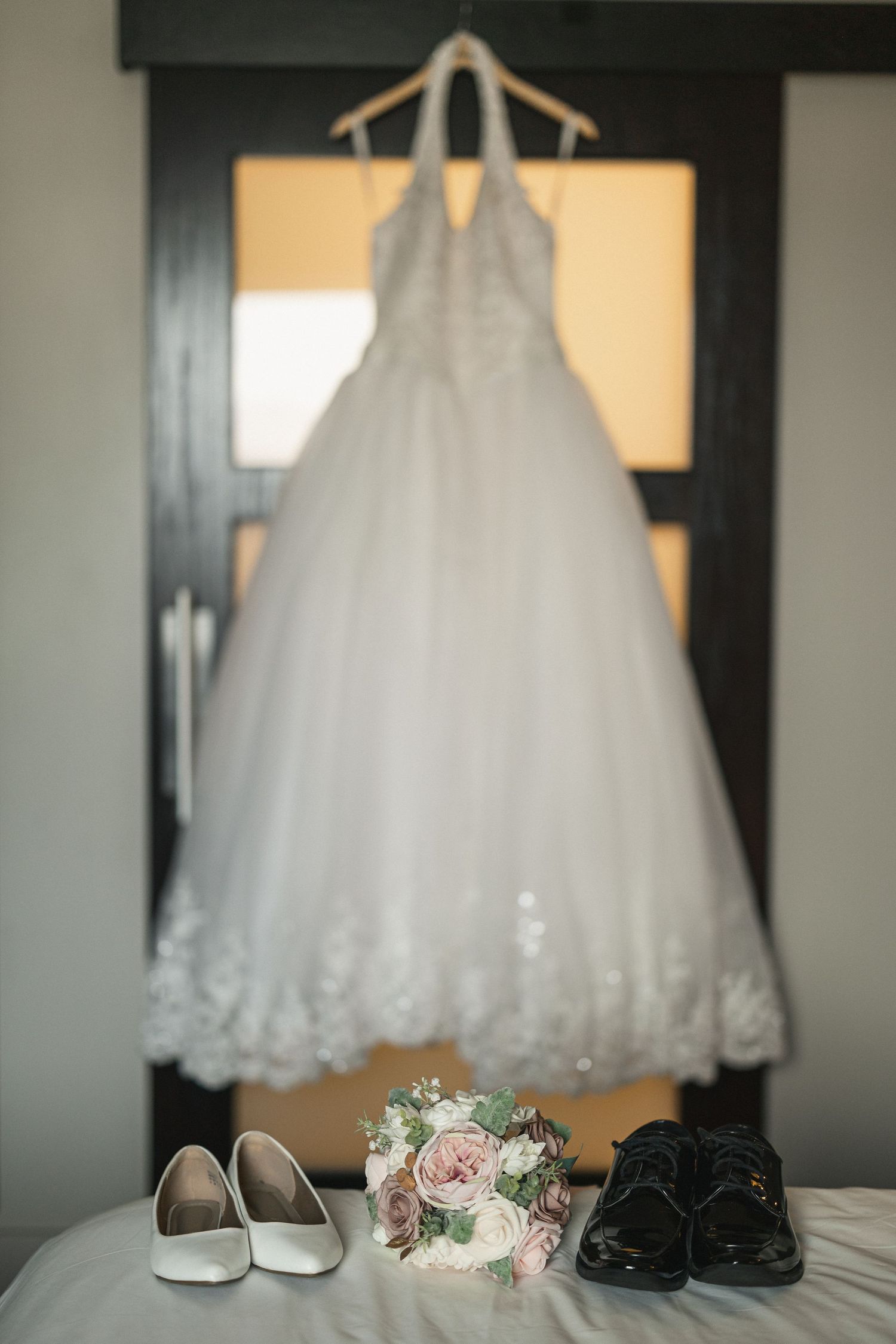 A white wedding dress hangs above bridal shoes and bouquet with pink roses on display.