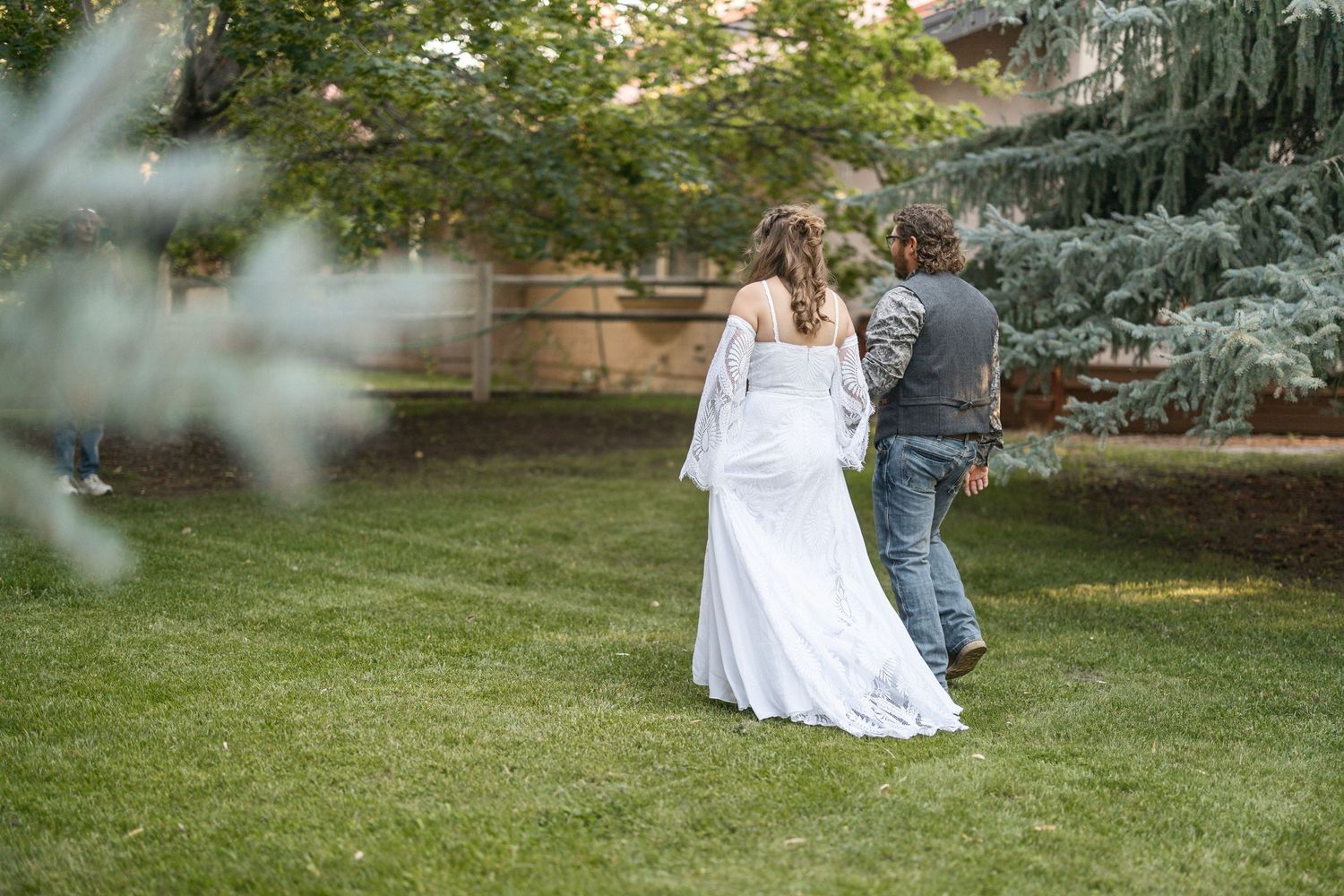 A couple in wedding attire sits together on a grassy lawn surrounded by trees in a garden setting.