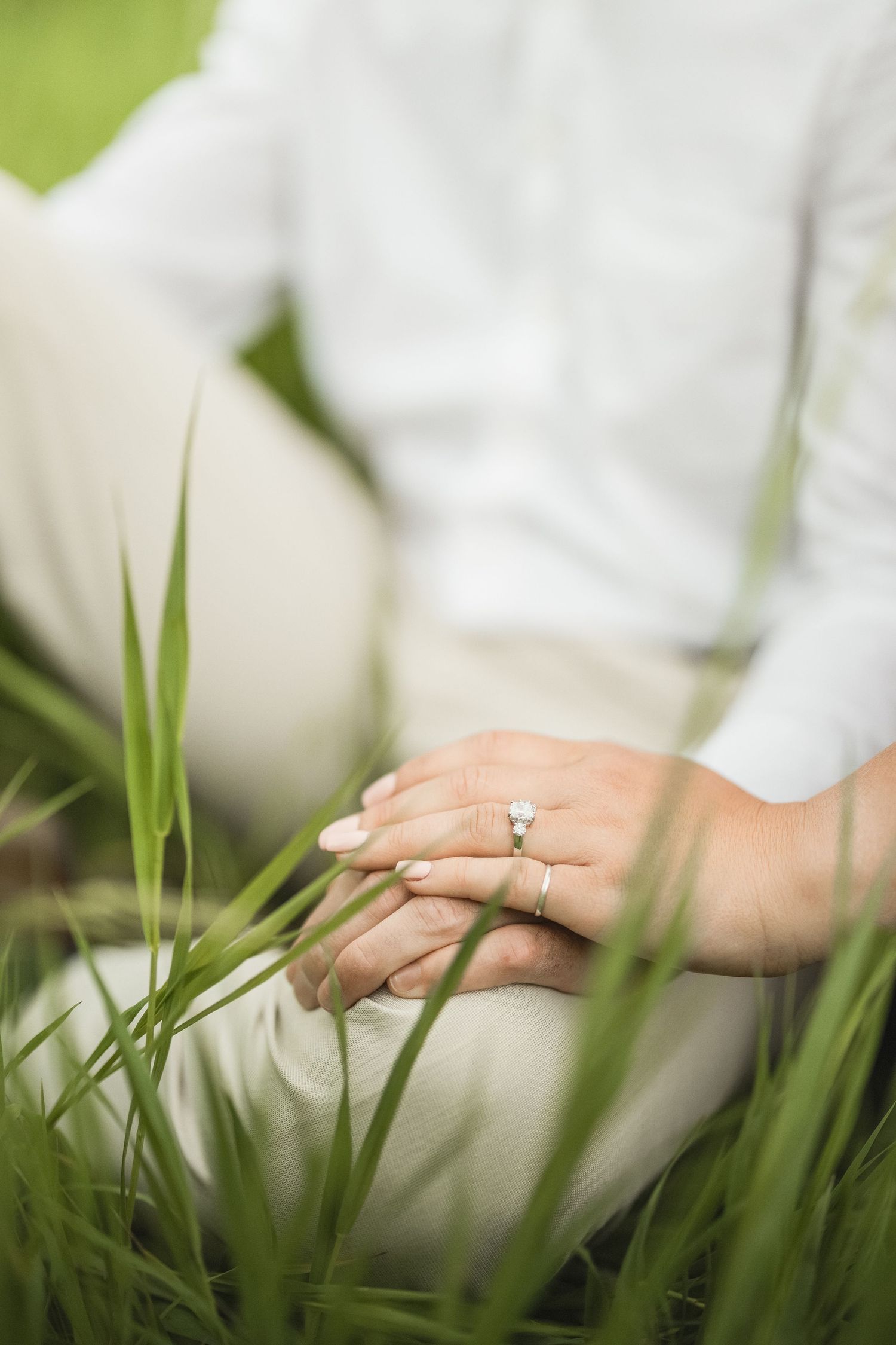 A delicate engagement ring sparkles on a hand resting in tall green grass on a sunny day.