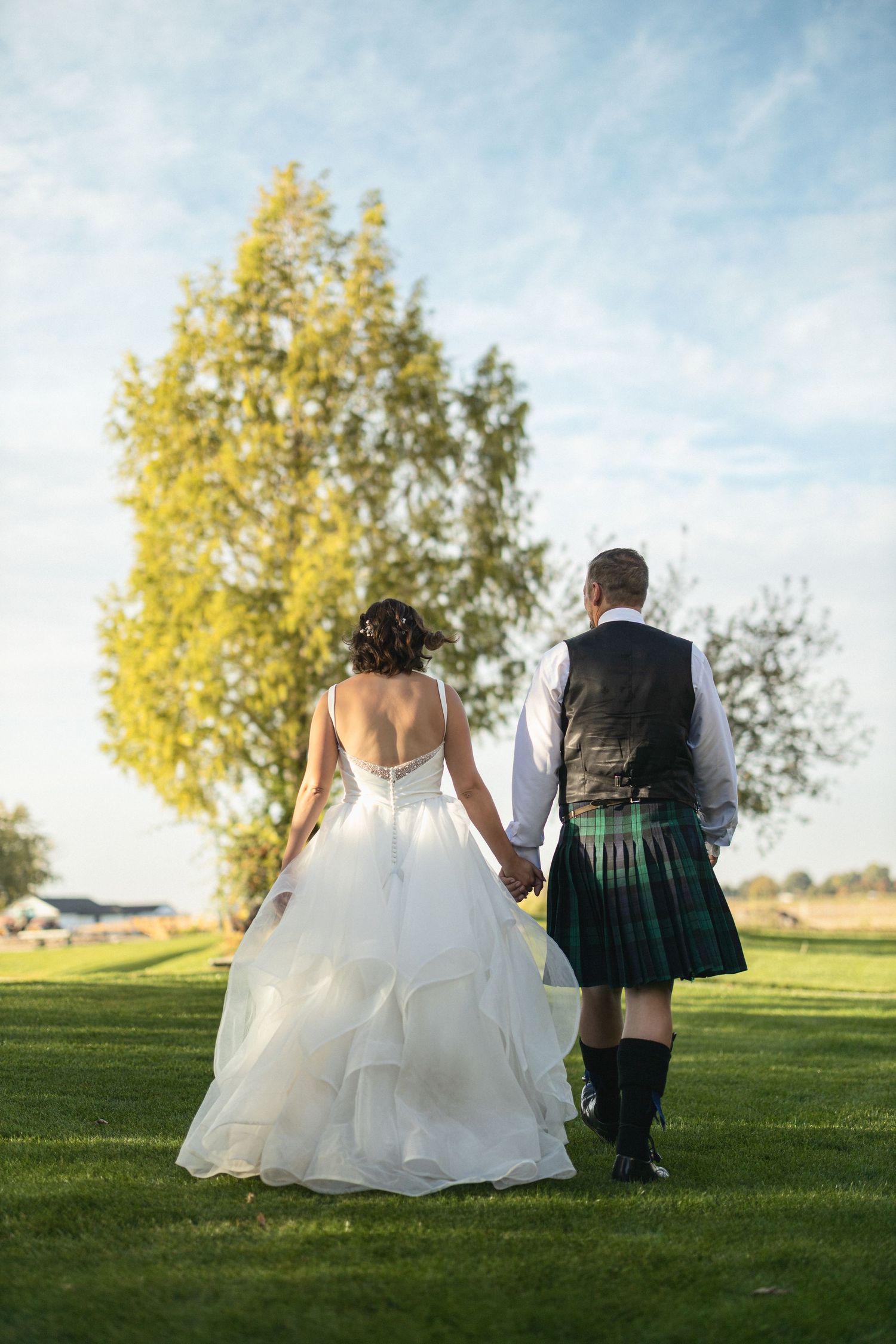 A couple walks hand in hand across a grassy field with a white church spire visible in the background.