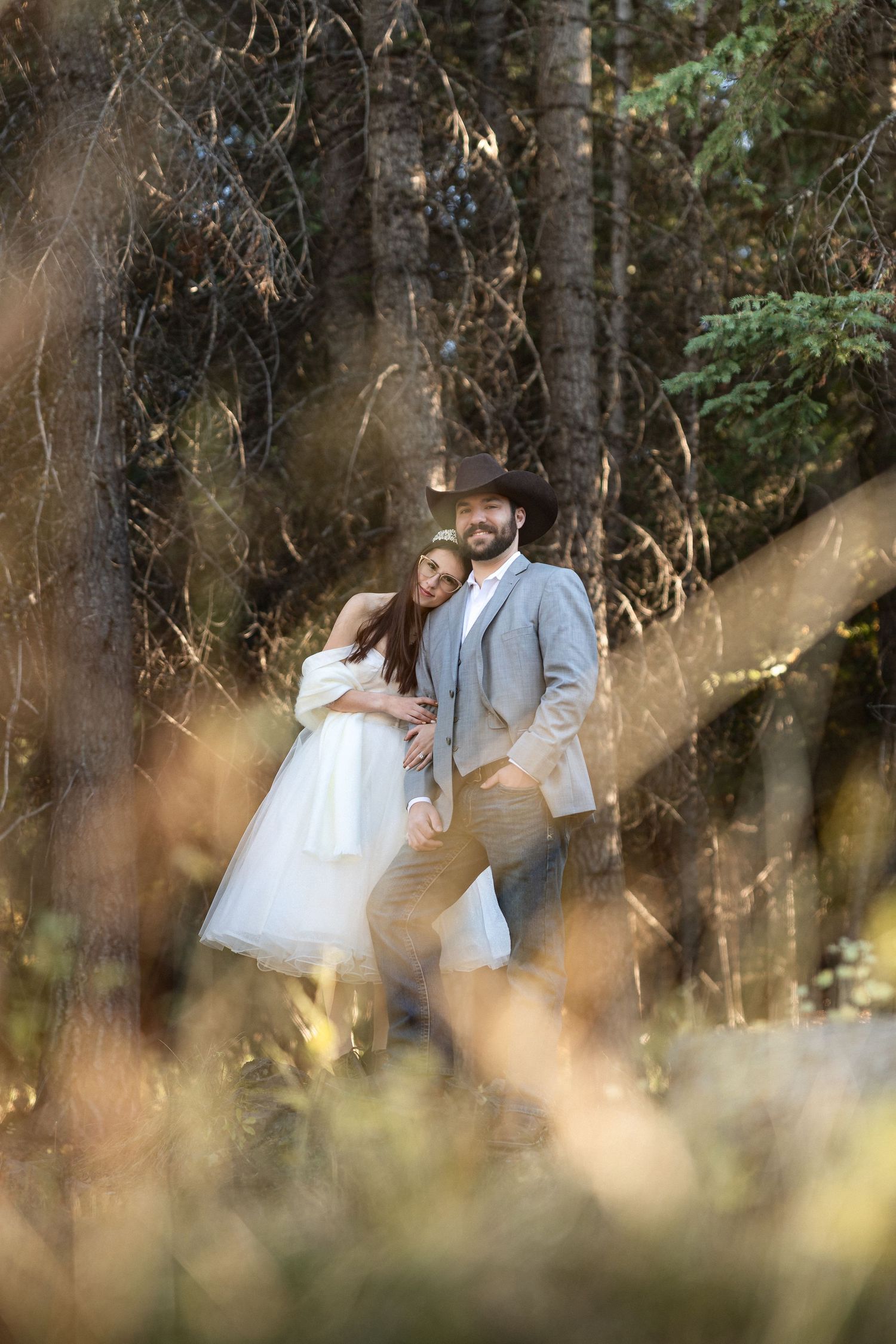 A wedding couple poses in a dreamy forest setting with golden sunlight filtering through the trees.