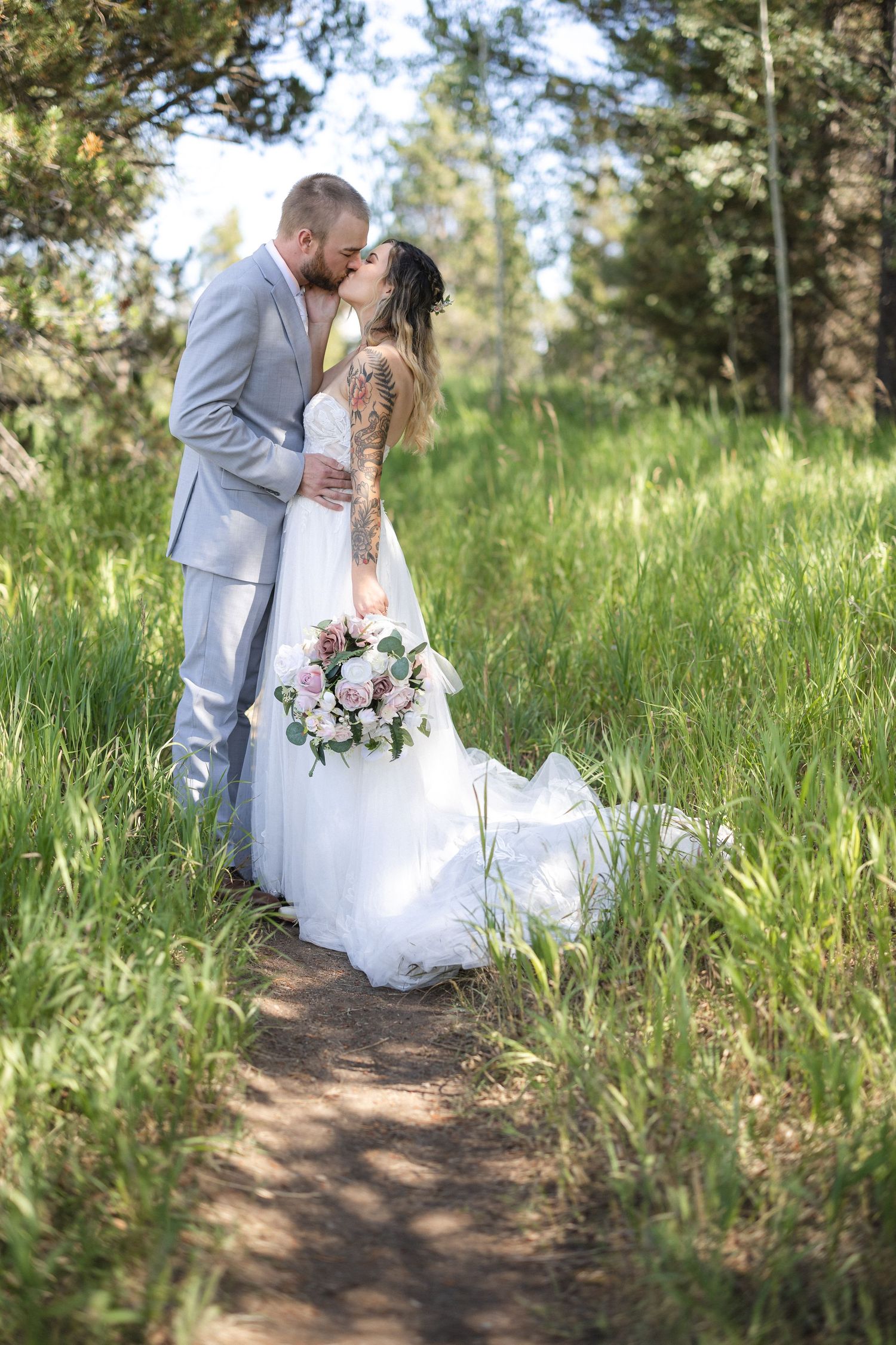 A couple embraces in a grassy outdoor setting with pine trees in the background.