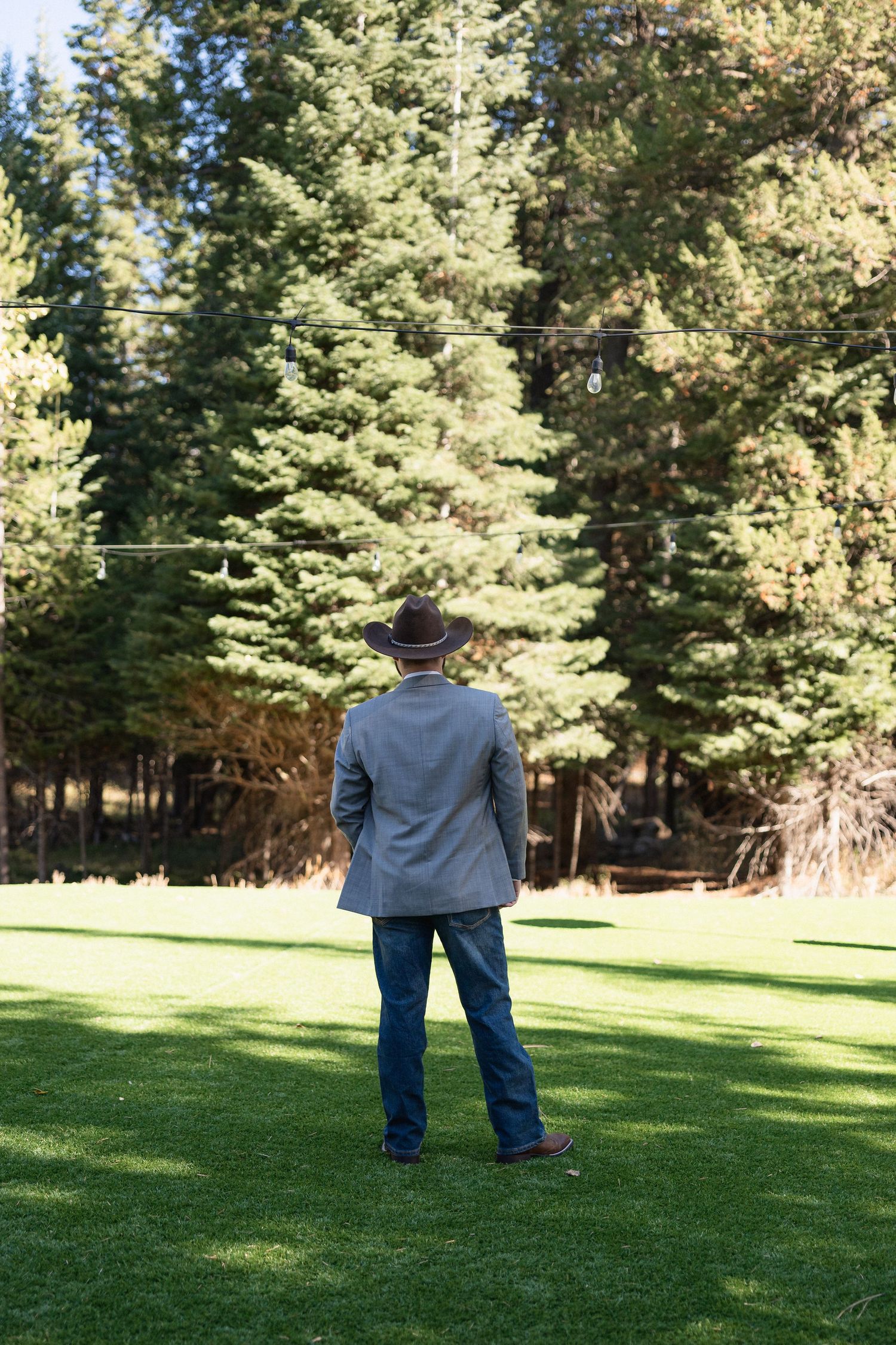 Person in grey suit and hat standing on grass facing evergreen trees.