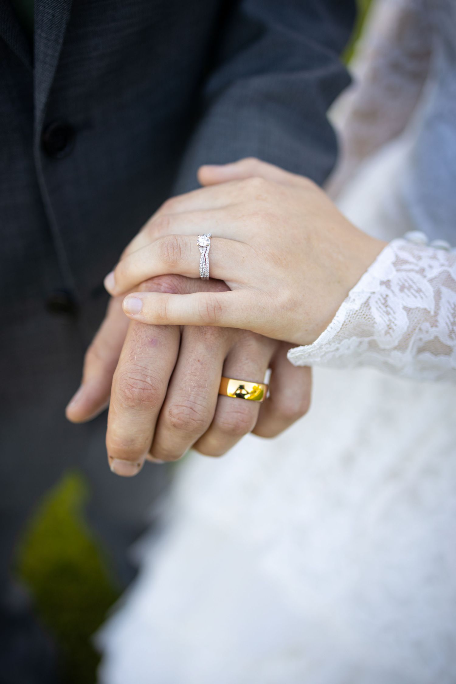 Close-up of bride and groom's hands with wedding rings against white lace dress and dark suit.