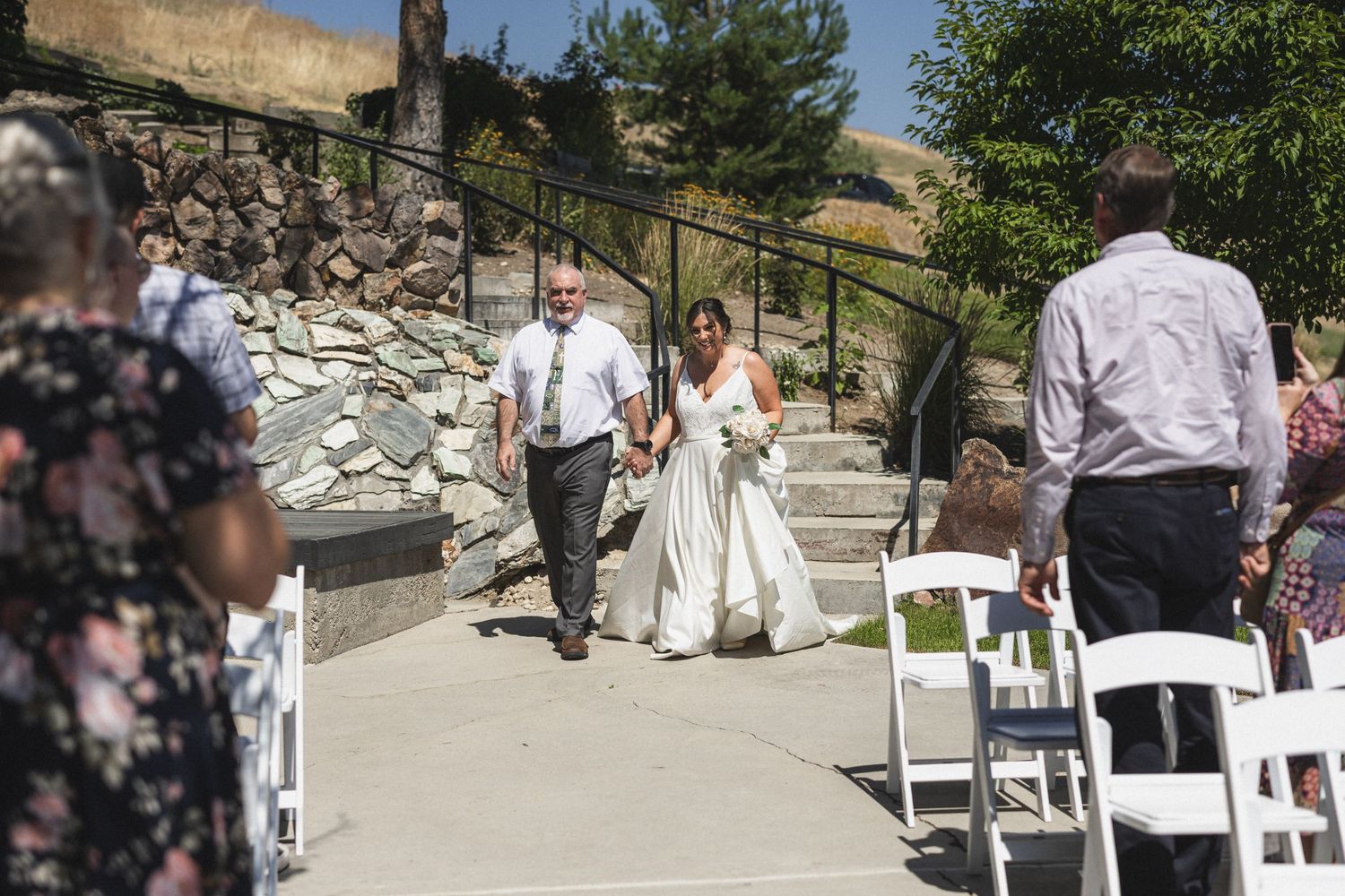 A bride in white walks up stone steps during an outdoor wedding ceremony.