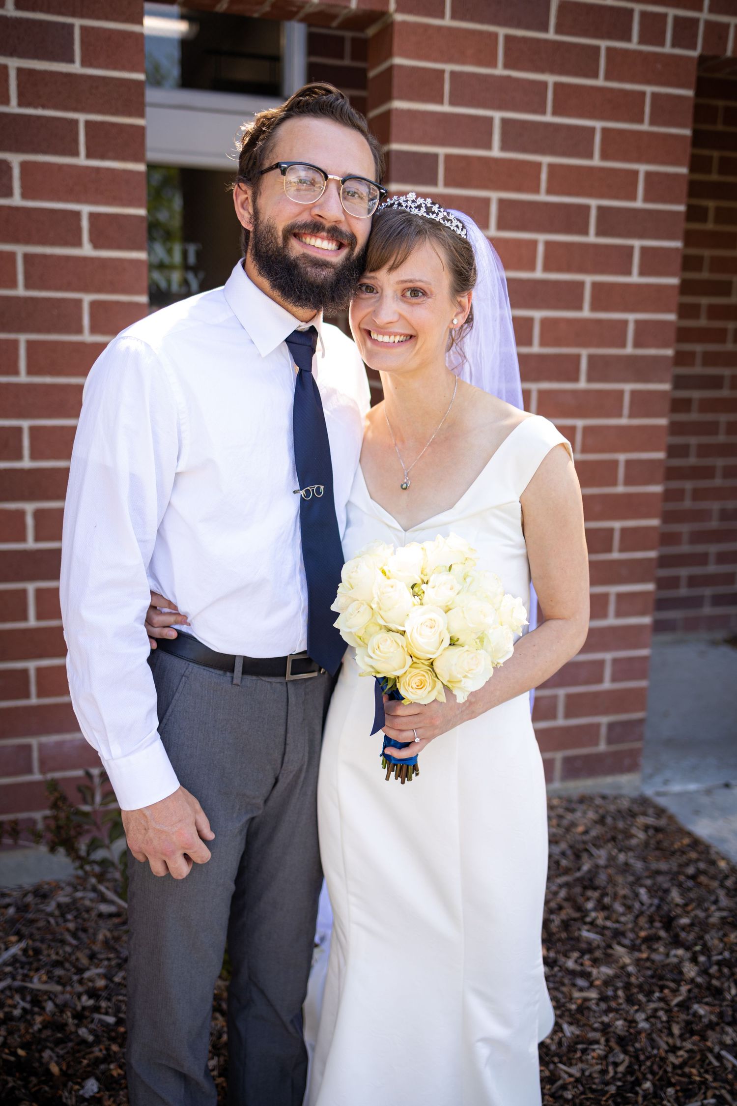 A happy couple poses together against a brick wall on their wedding day, with the bride holding yellow flowers.