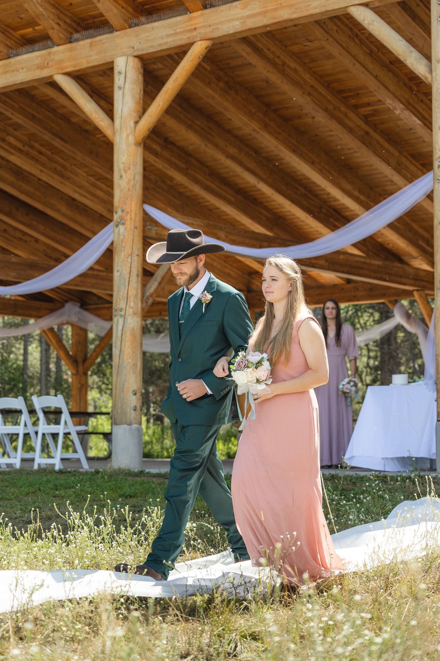 Wedding party members in formal attire walking under wooden pavilion with draped fabric decor.