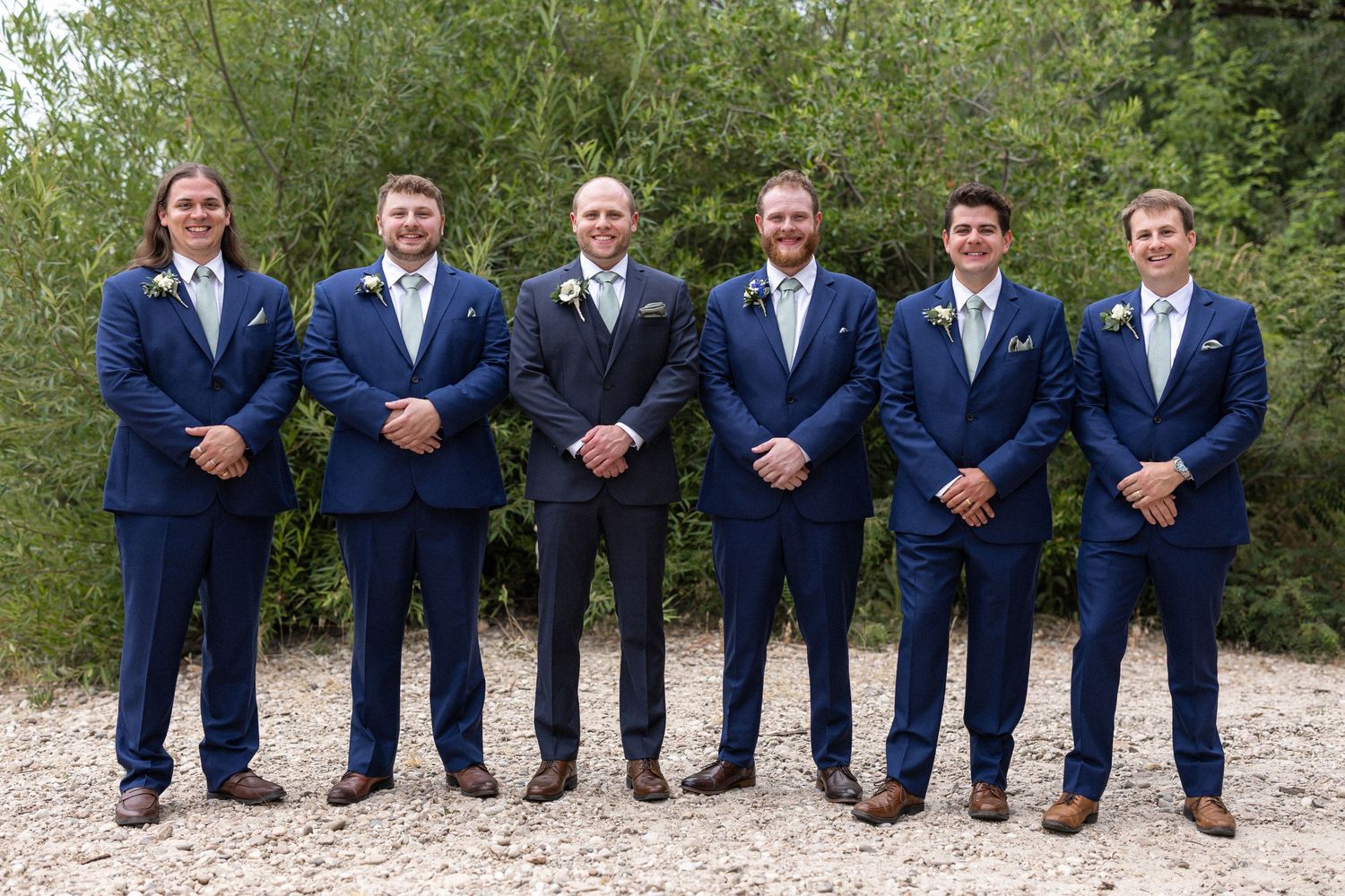 Groomsmen in navy blue suits pose formally outdoors on gravel path.