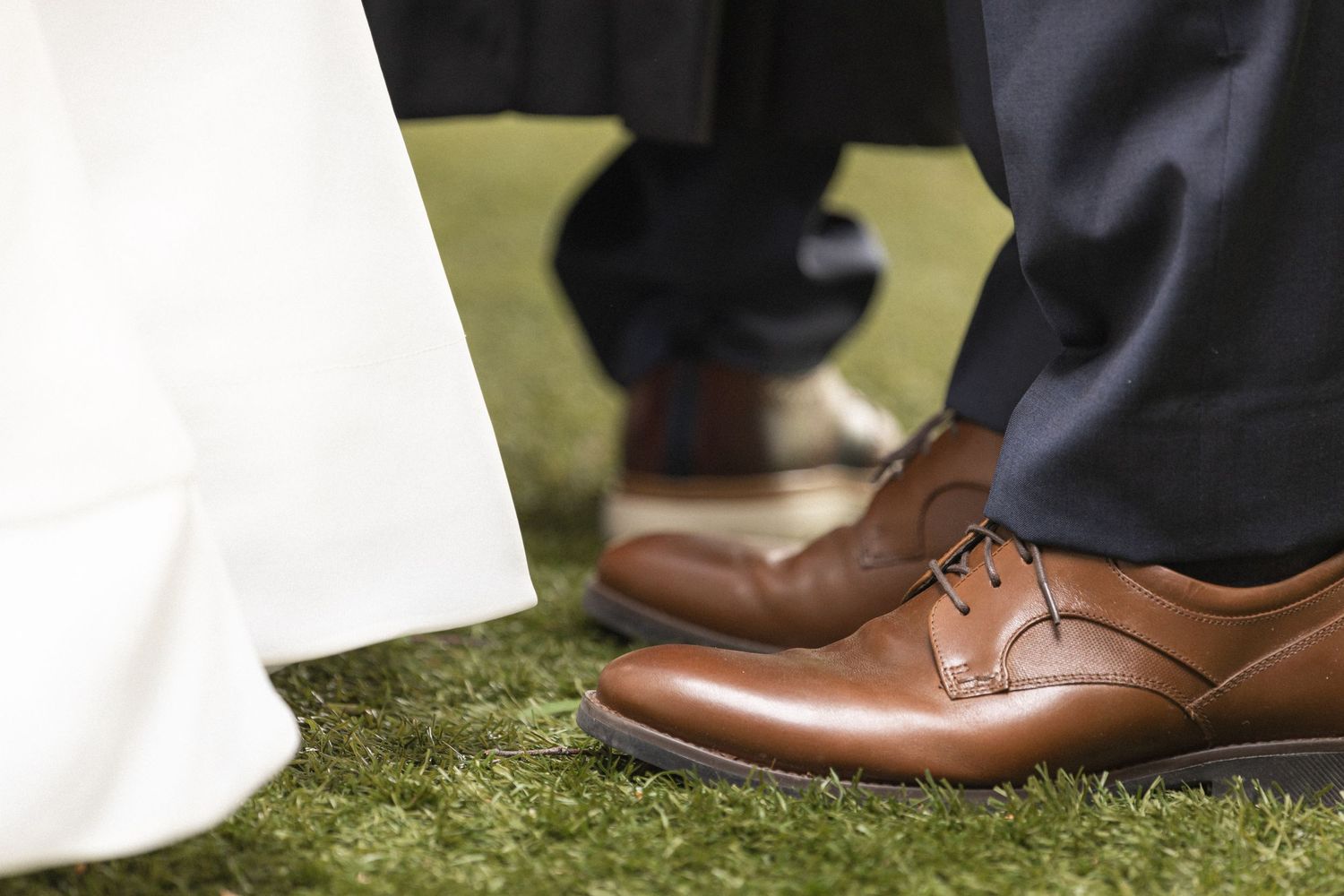 Close-up shot of brown leather dress shoes and white wedding dress hem on grass.