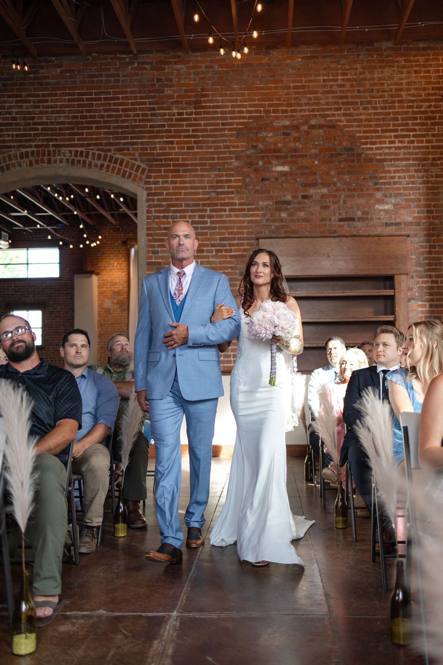 Wedding processional in industrial brick venue with string lights and seated guests.