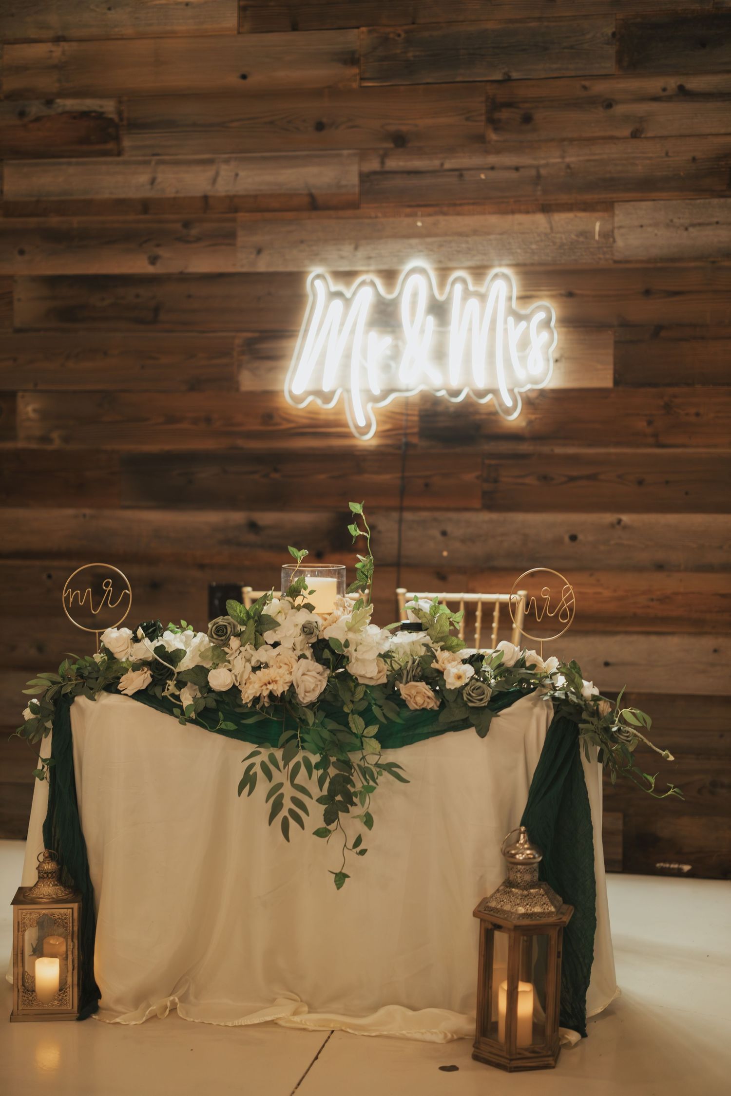 Wedding reception table with neon sign, white floral arrangements and lanterns against wooden wall.