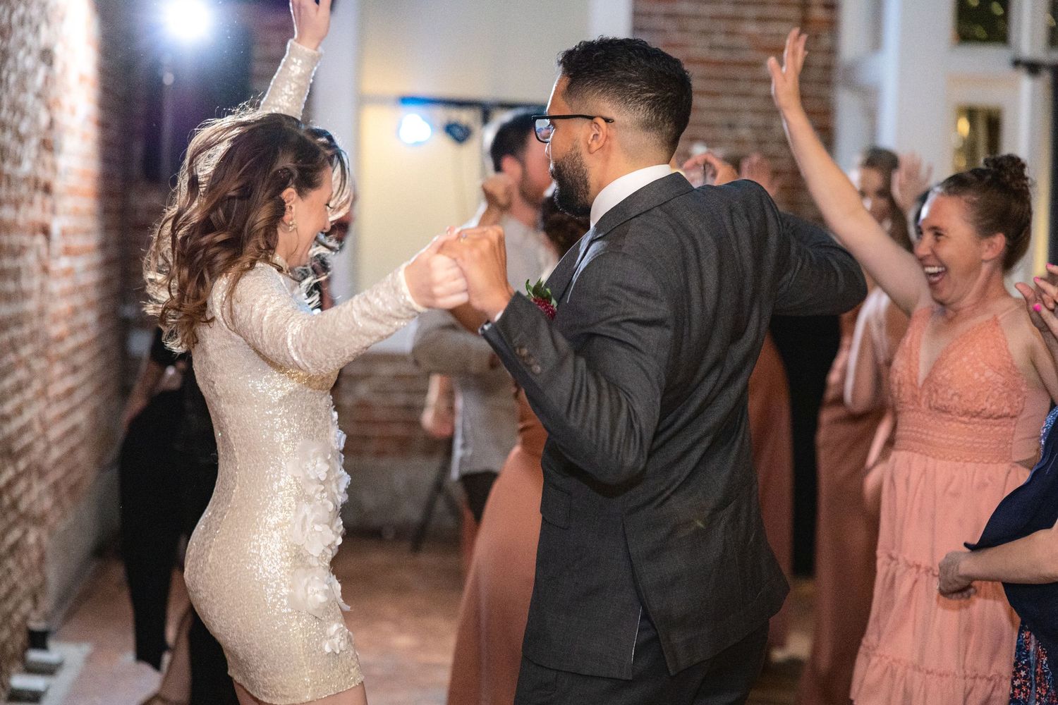 Wedding celebration with guests dancing in formal attire at an indoor brick venue.