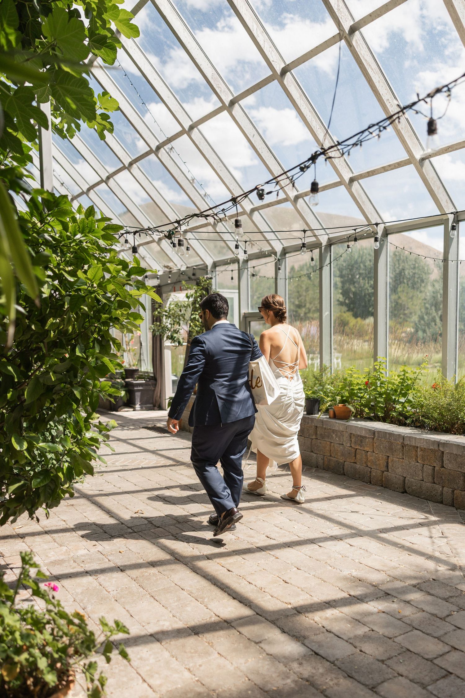 Couple exploring a sunlit greenhouse with glass panels and stone walls on a bright day.