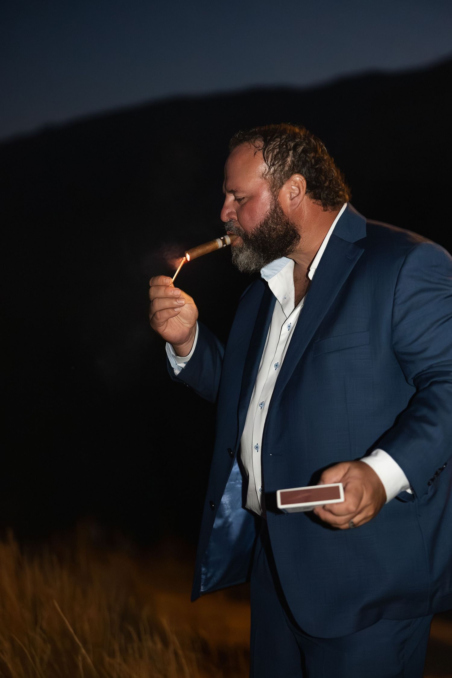 Wedding party member in navy suit enjoying a cigar during outdoor evening celebrations.