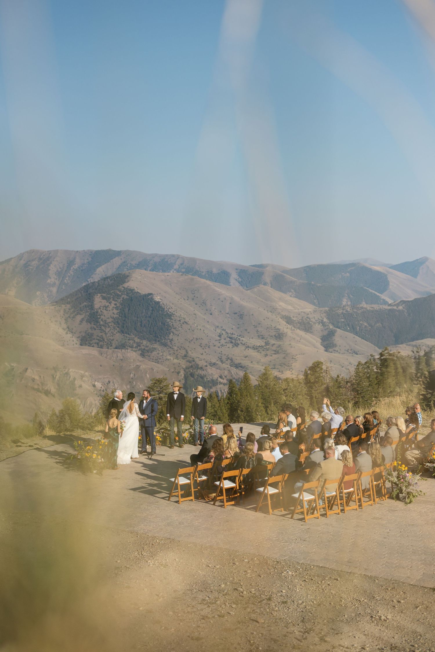 A dreamy, sun-flared photograph of a mountain wedding ceremony with guests seated on wooden chairs.