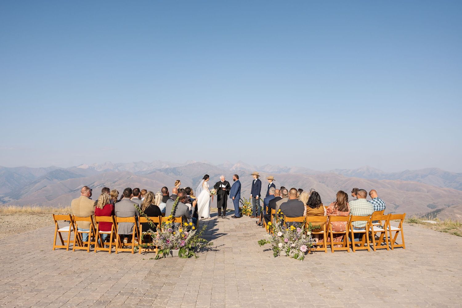 A panoramic view of a mountaintop wedding ceremony with wooden chairs arranged in rows under clear blue skies.