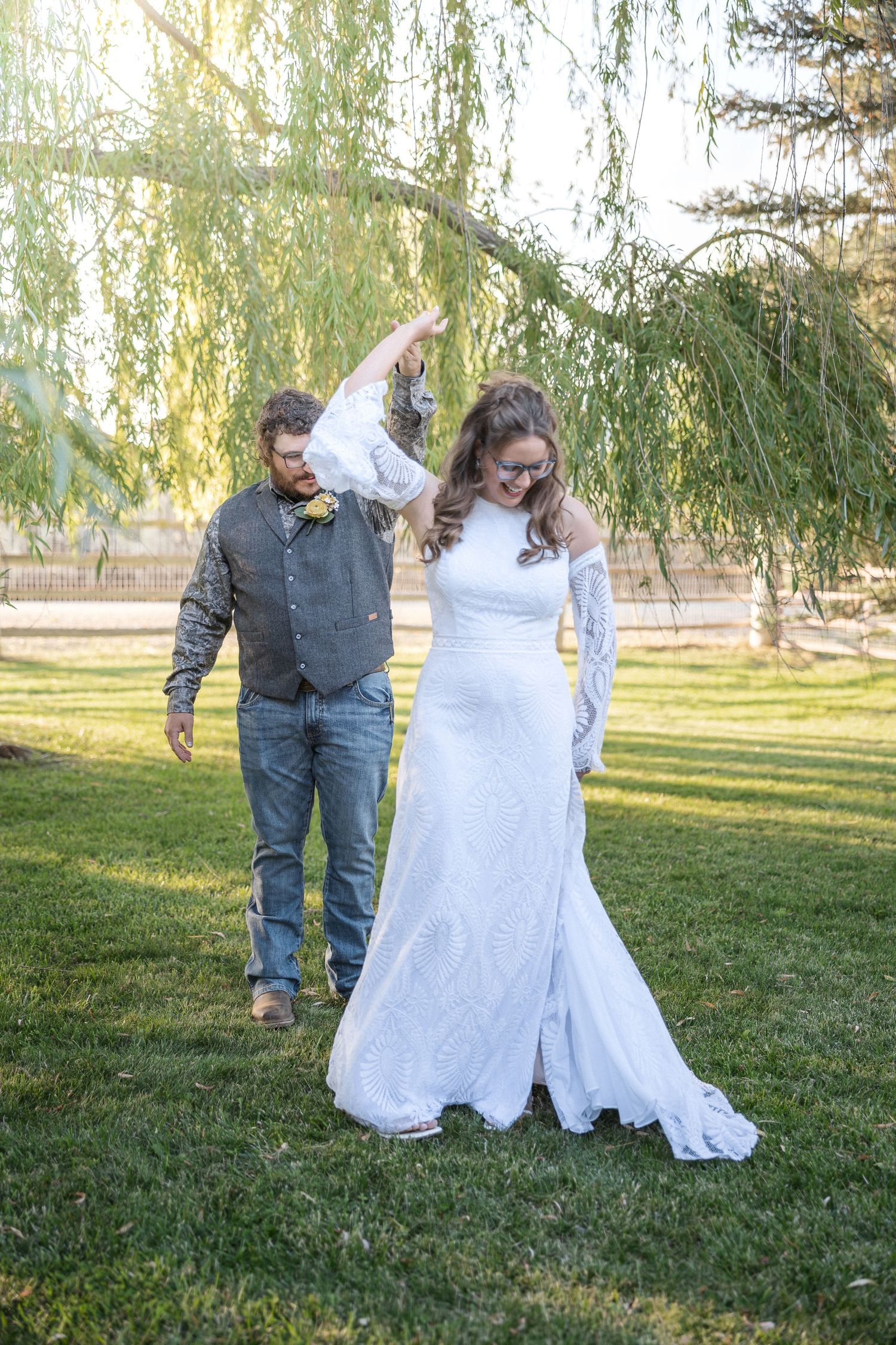 Wedding couple dancing together on grass under weeping willow trees at sunset.