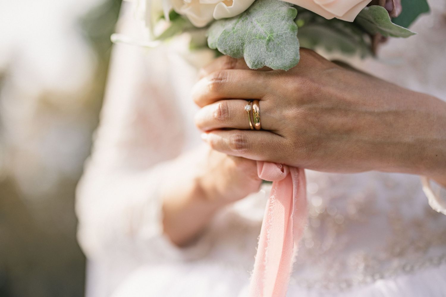 Close-up of a bride's hand wearing a wedding ring while holding a bouquet with soft pink details.