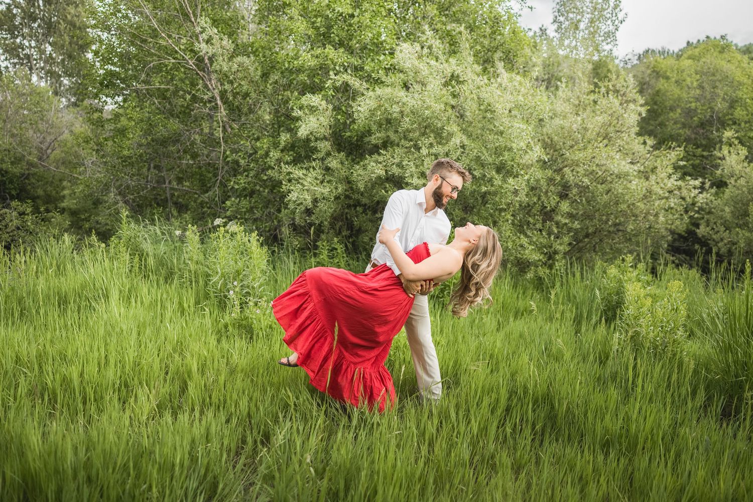 A couple in formal attire embraces romantically in a lush green meadow on a sunny summer day.