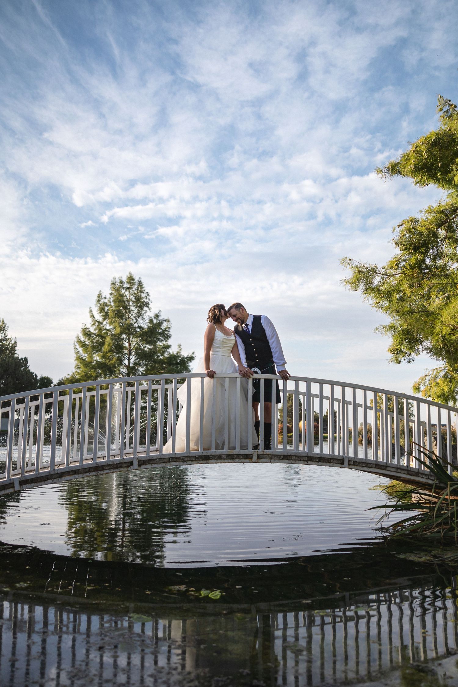 A romantic moment captured on a white bridge over still water with wispy clouds overhead.
