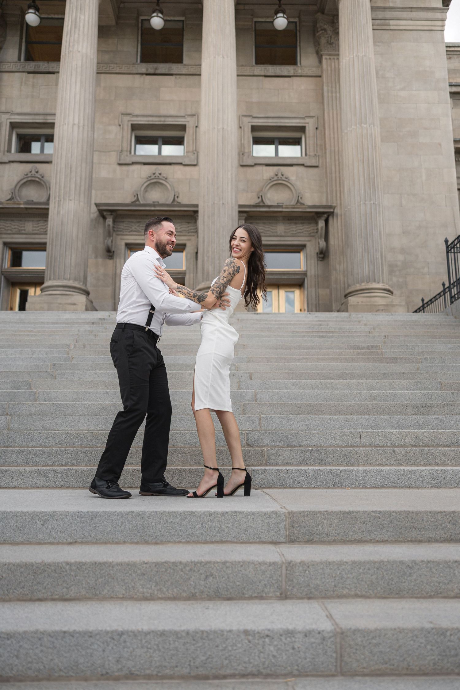 Couple in black and white attire poses playfully on grand stone steps of historic building.