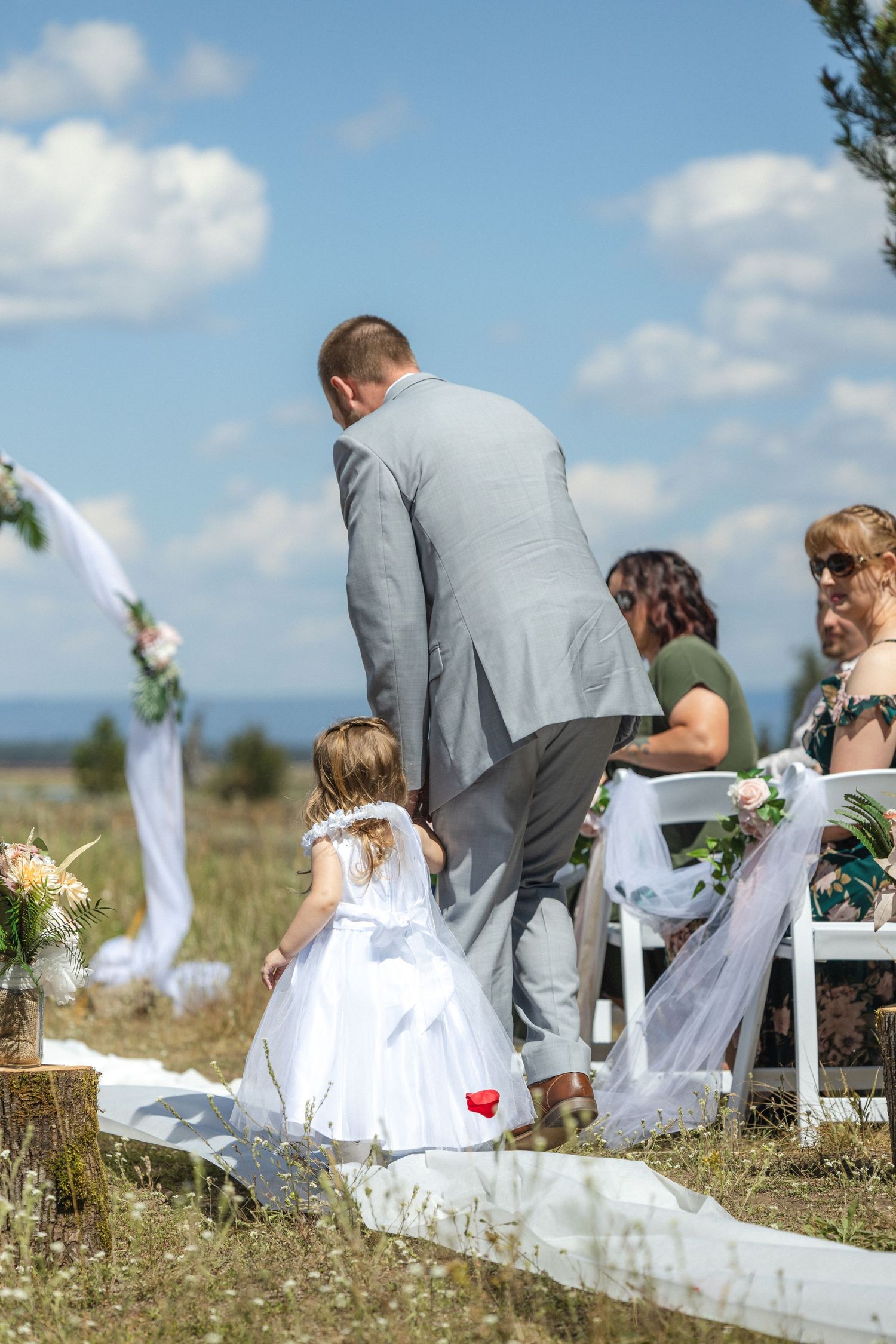A person in grey suit walking with small child in white dress down white aisle runner at outdoor wedding.