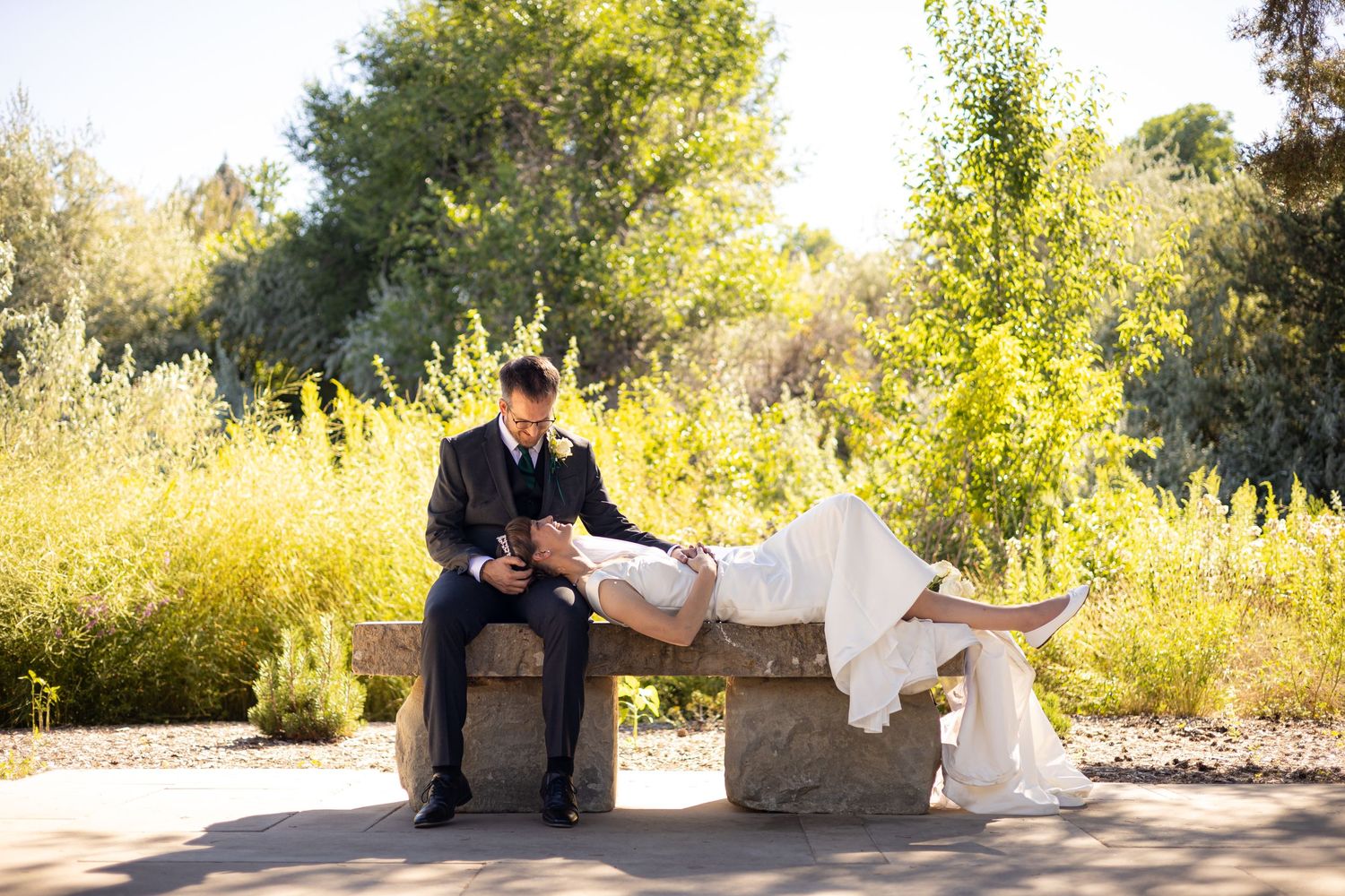 Wedding couple relaxes on stone bench surrounded by golden sunlit foliage.