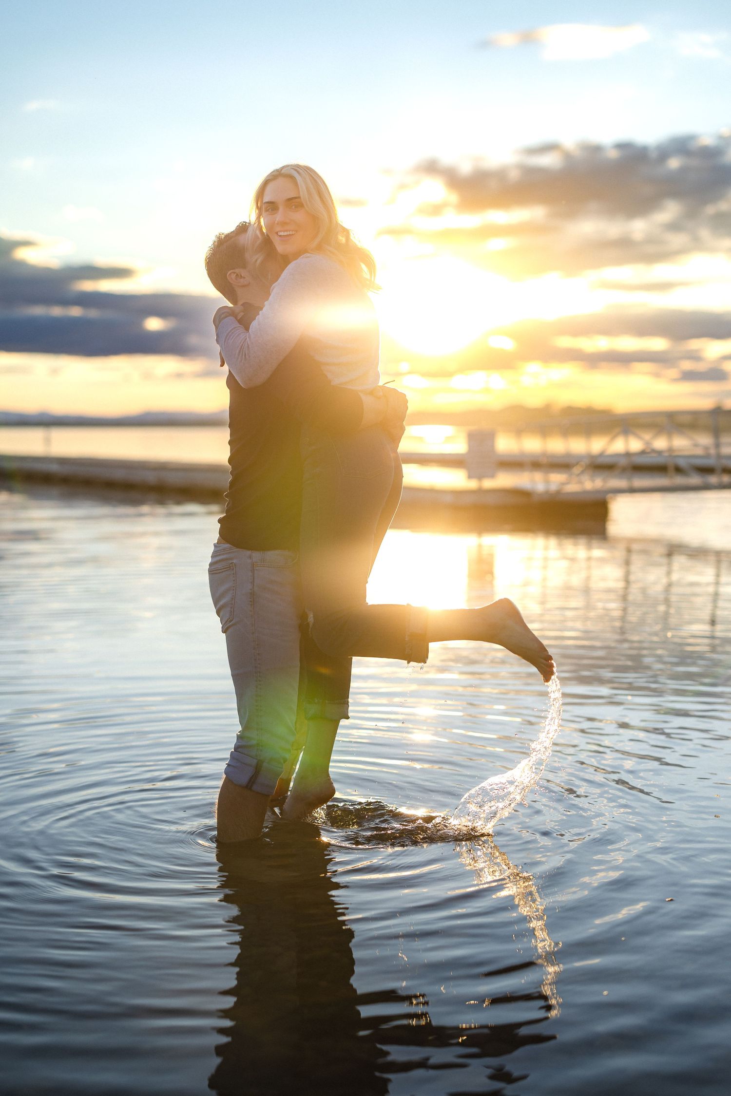 Joyful couple embracing in water with dramatic sunset and marina in background.