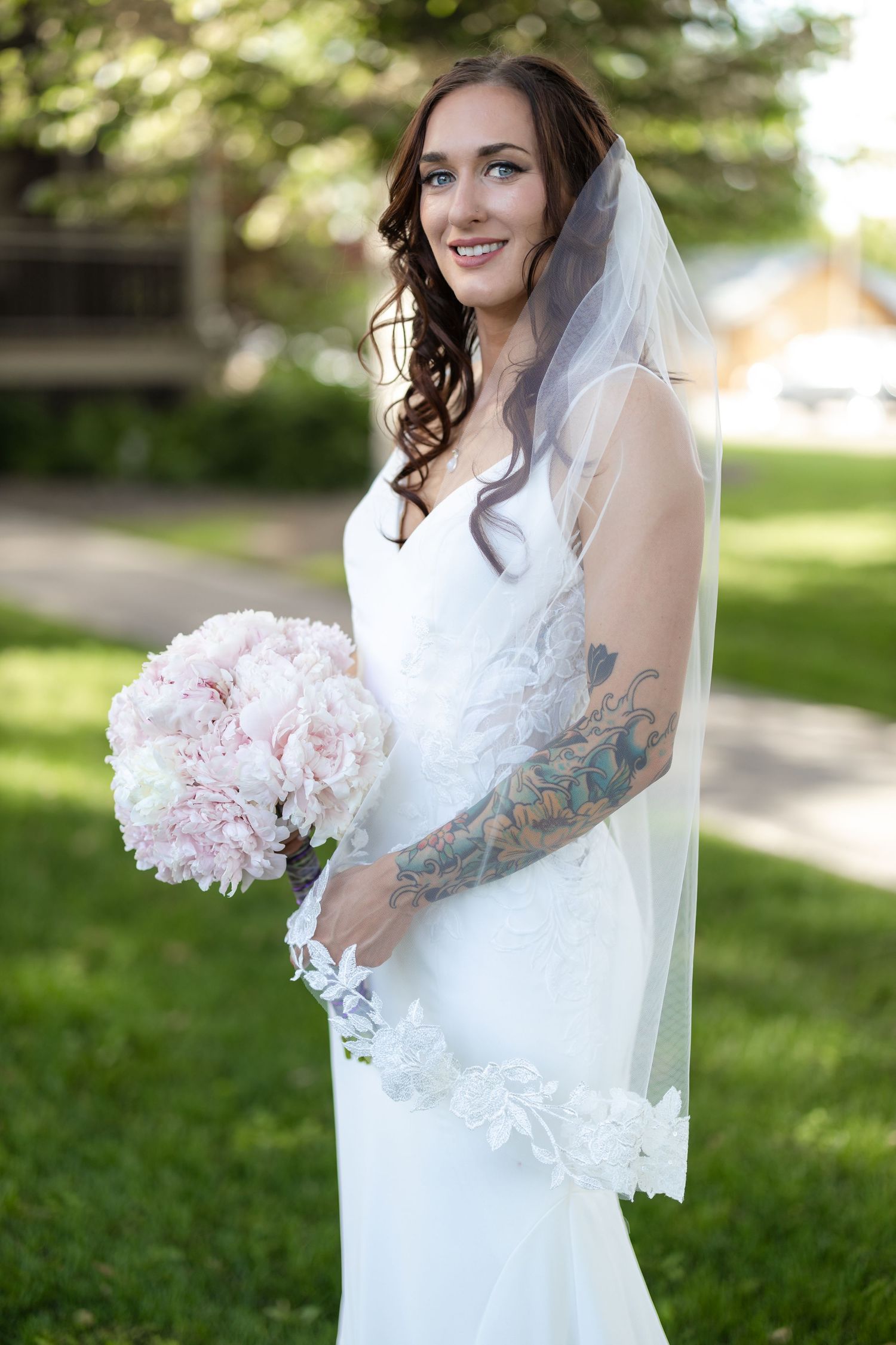 Bride in white dress with tattoo sleeve holds pink bouquet while wearing lace-trimmed veil.