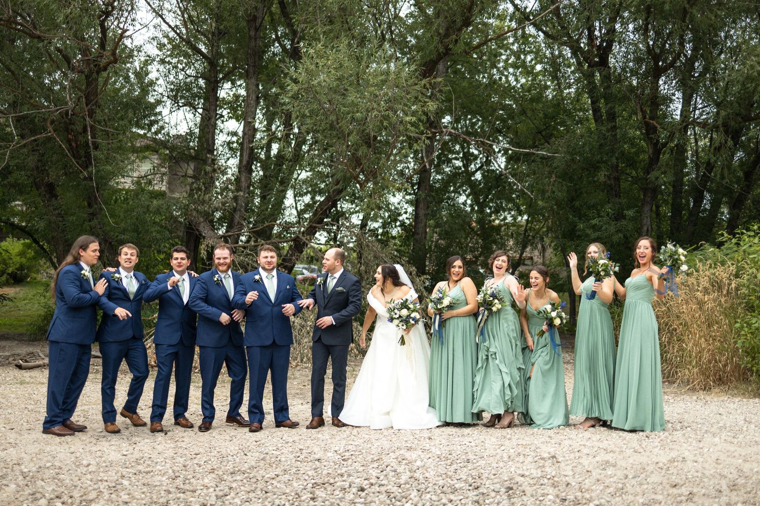 Wedding party stands on gravel path with bridesmaids in sage green dresses and groomsmen in navy suits.