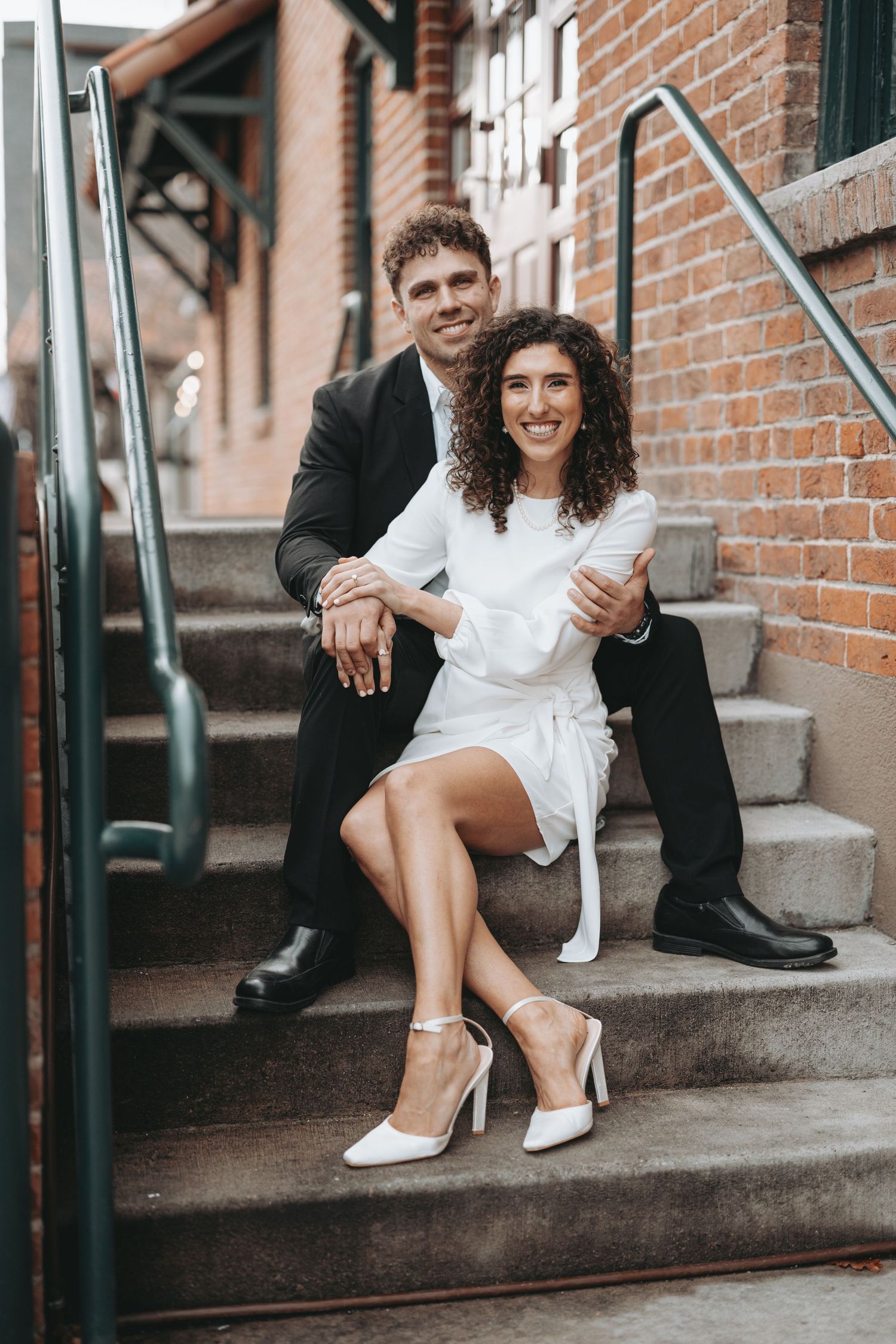 A couple in formal attire posed on concrete steps with brick wall backdrop.