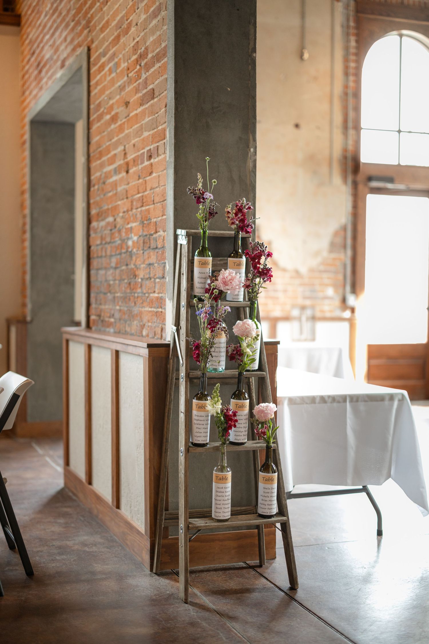Decorative ladder display with hanging glass bottles and flowers against brick wall.