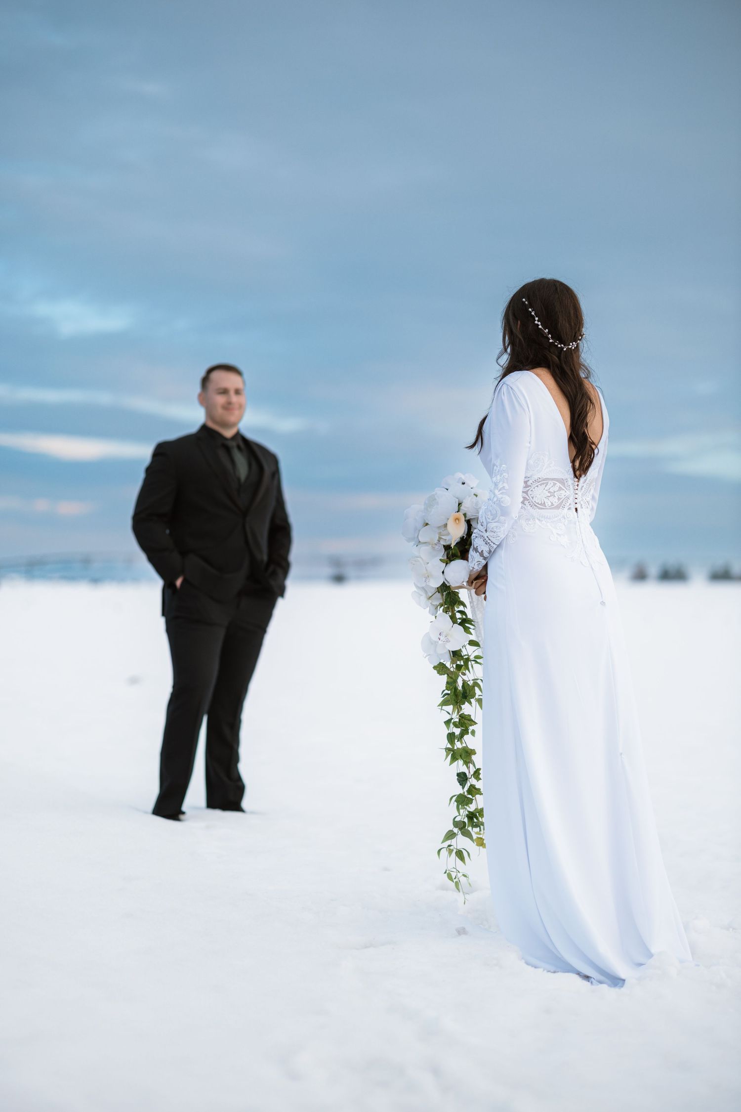Couple in winter wedding attire gazes at each other across snowy landscape with cloudy blue sky backdrop.