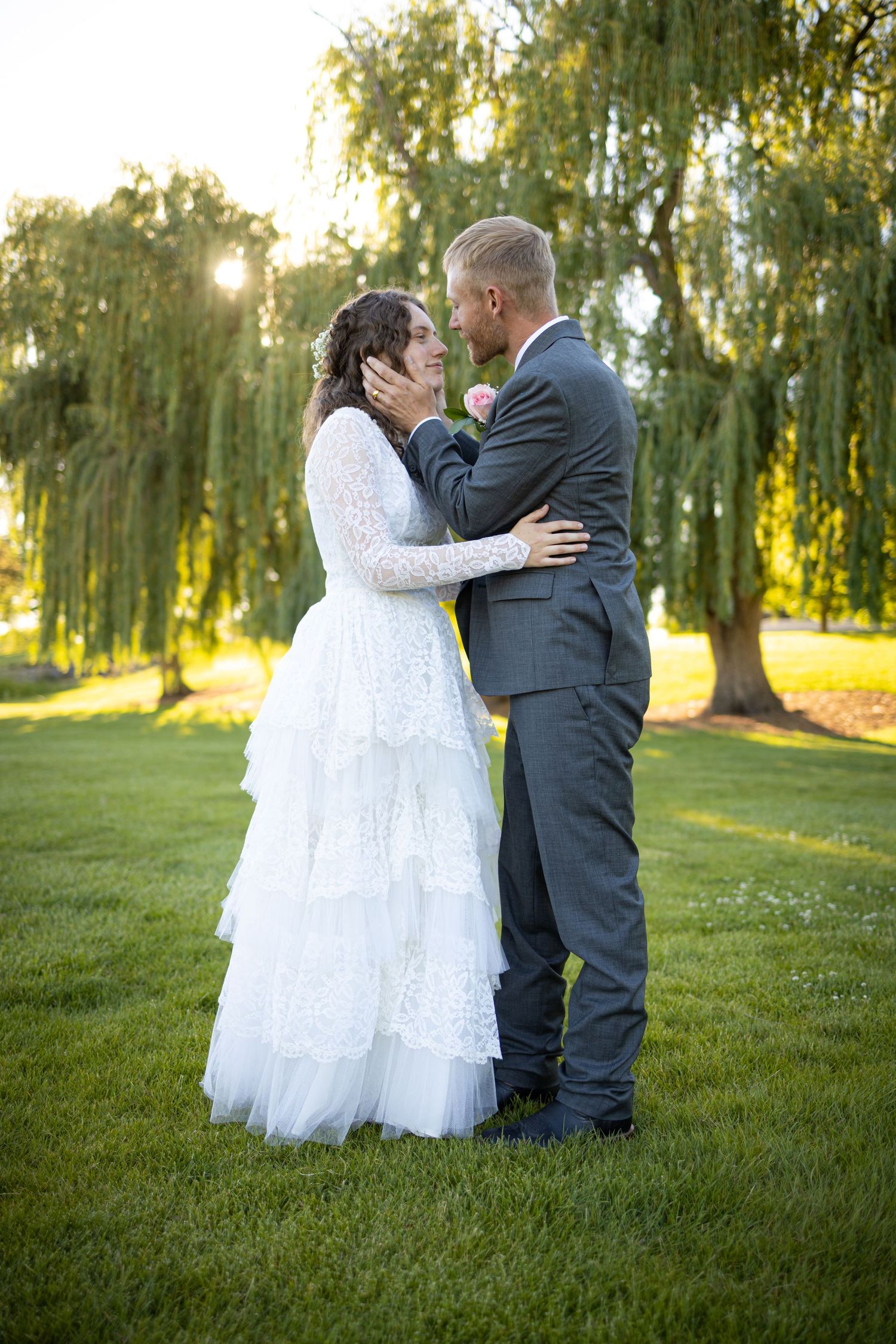 Tender embrace between couple in wedding attire with willow trees and sunset in background.