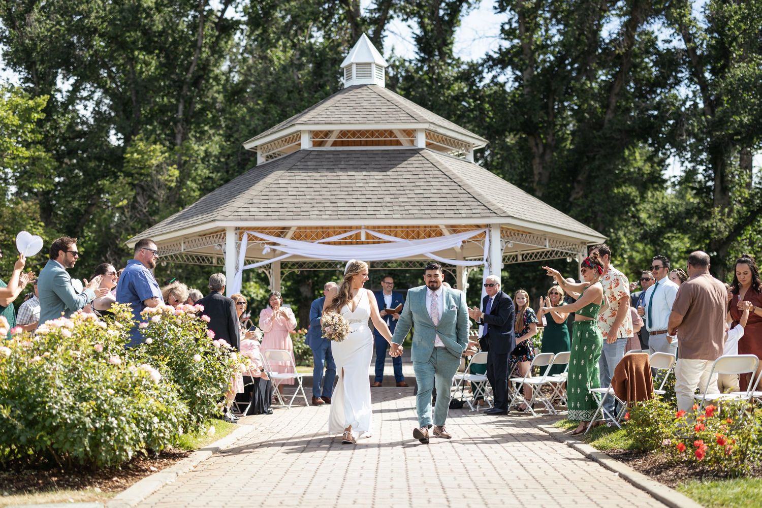 Outdoor garden wedding celebration with guests gathered around white gazebo.