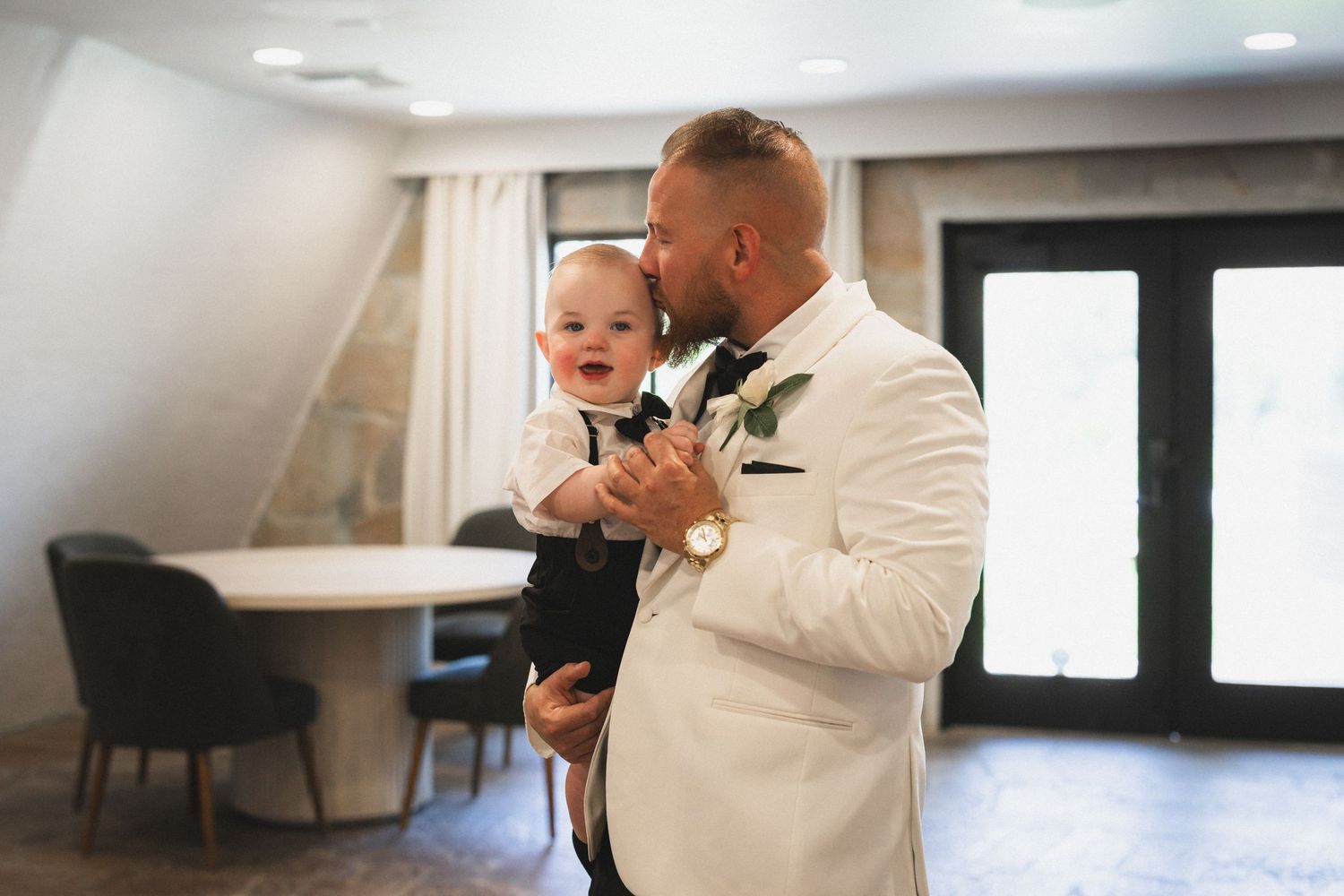 A person in a white tuxedo jacket holding a small baby in formal wear.