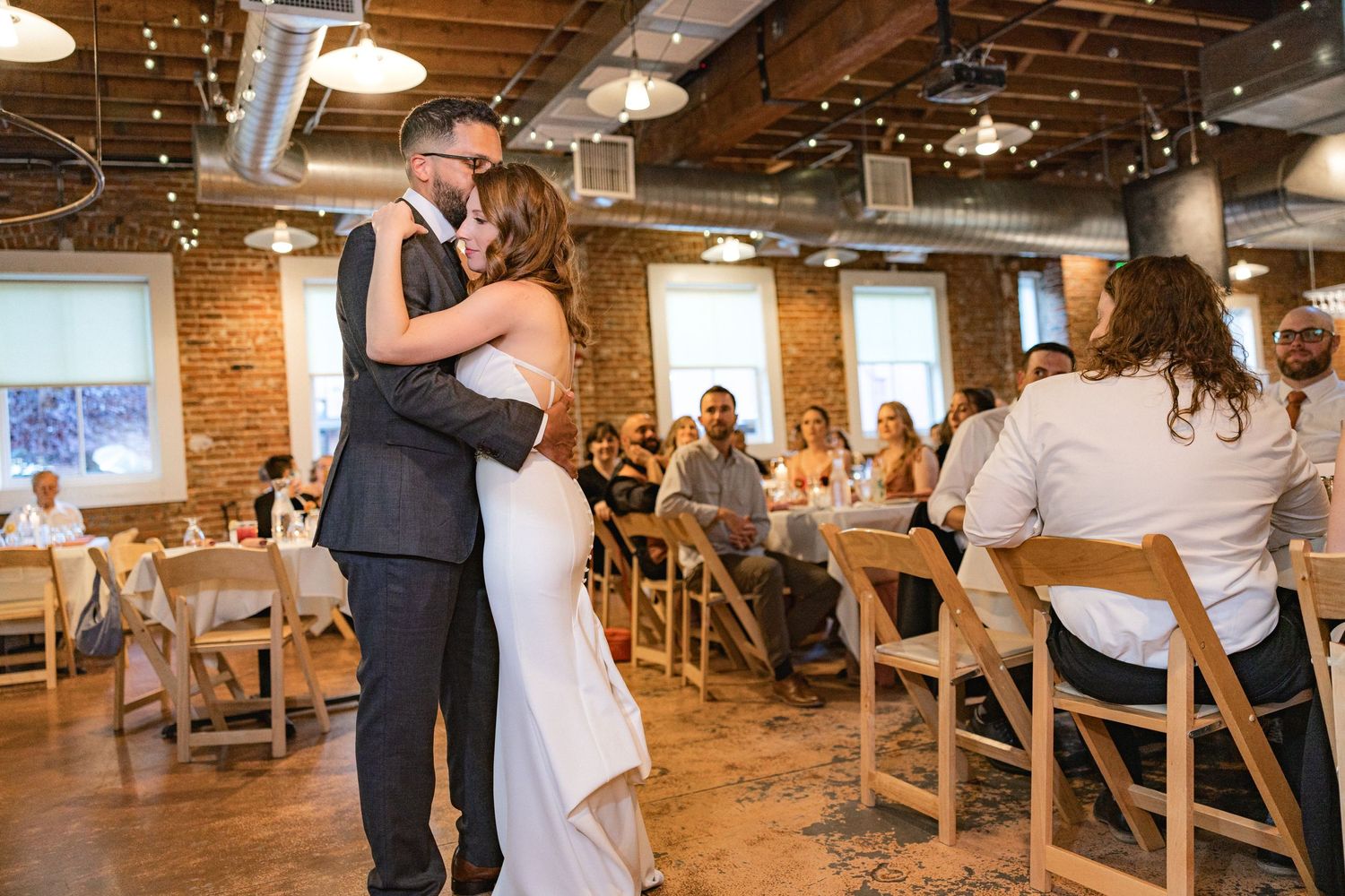 Newlyweds share intimate moment during reception in rustic brick venue with string lights.