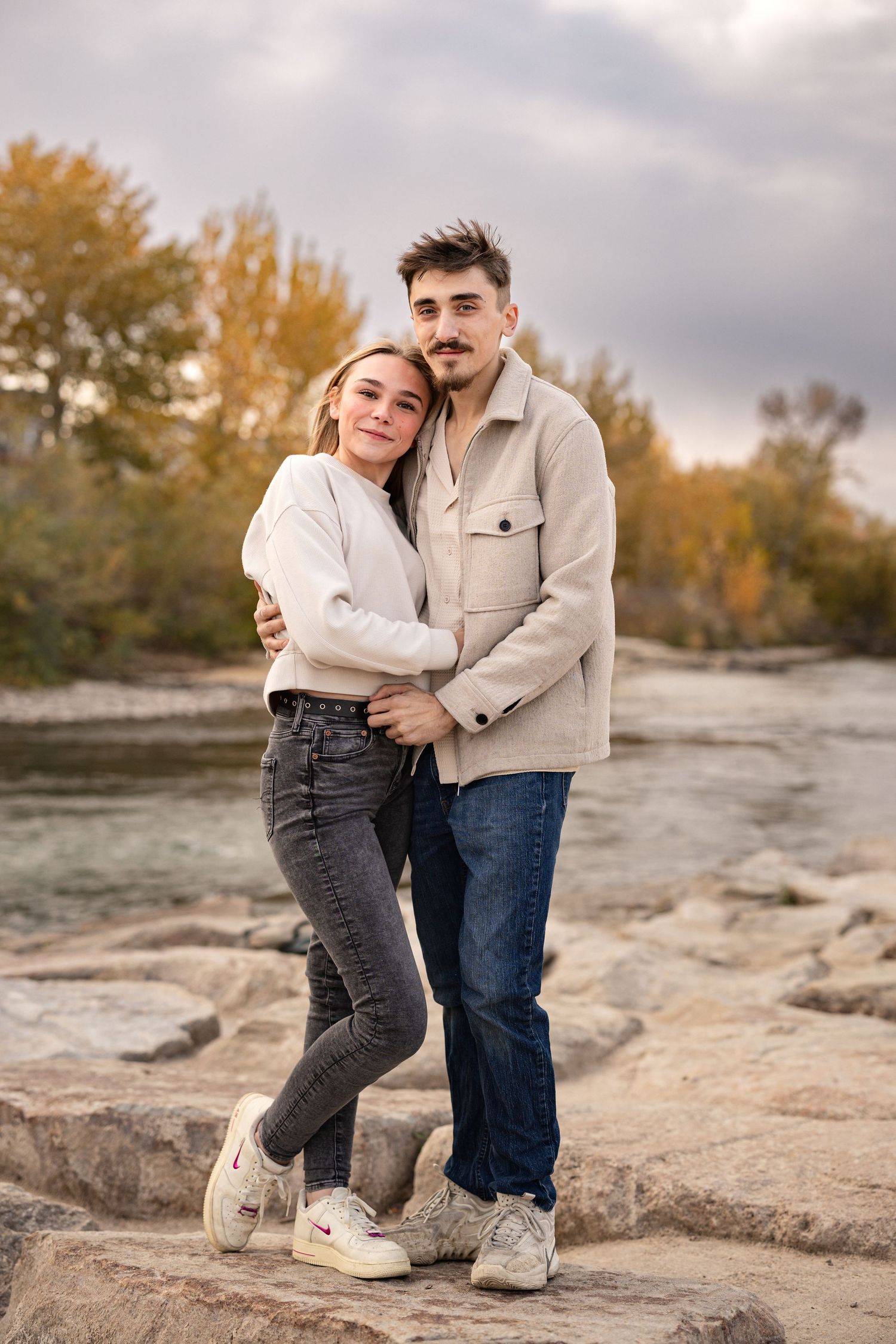 A couple shares a romantic moment on rocky terrain during autumn with warm golden light and trees in the background.