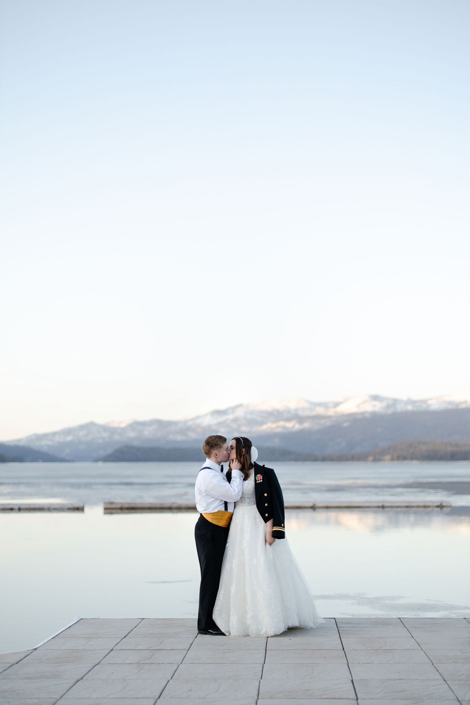 A couple shares an intimate moment by a serene lake with snow-capped mountains in the distance.
