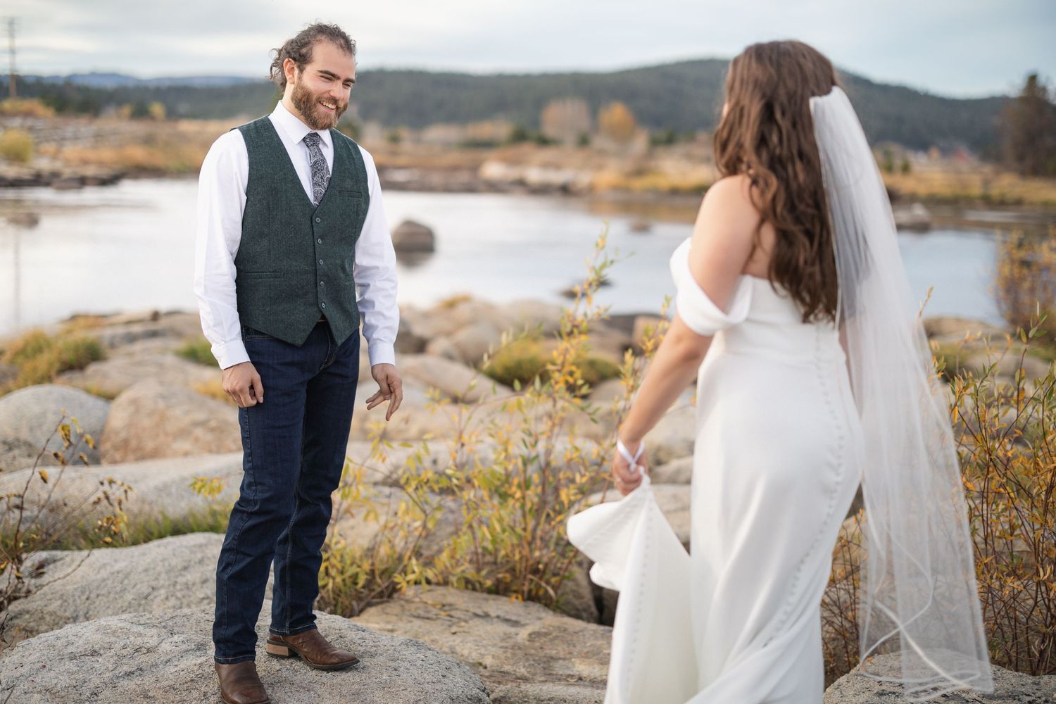 Wedding couple standing on rocky terrain with lake and mountains in background.