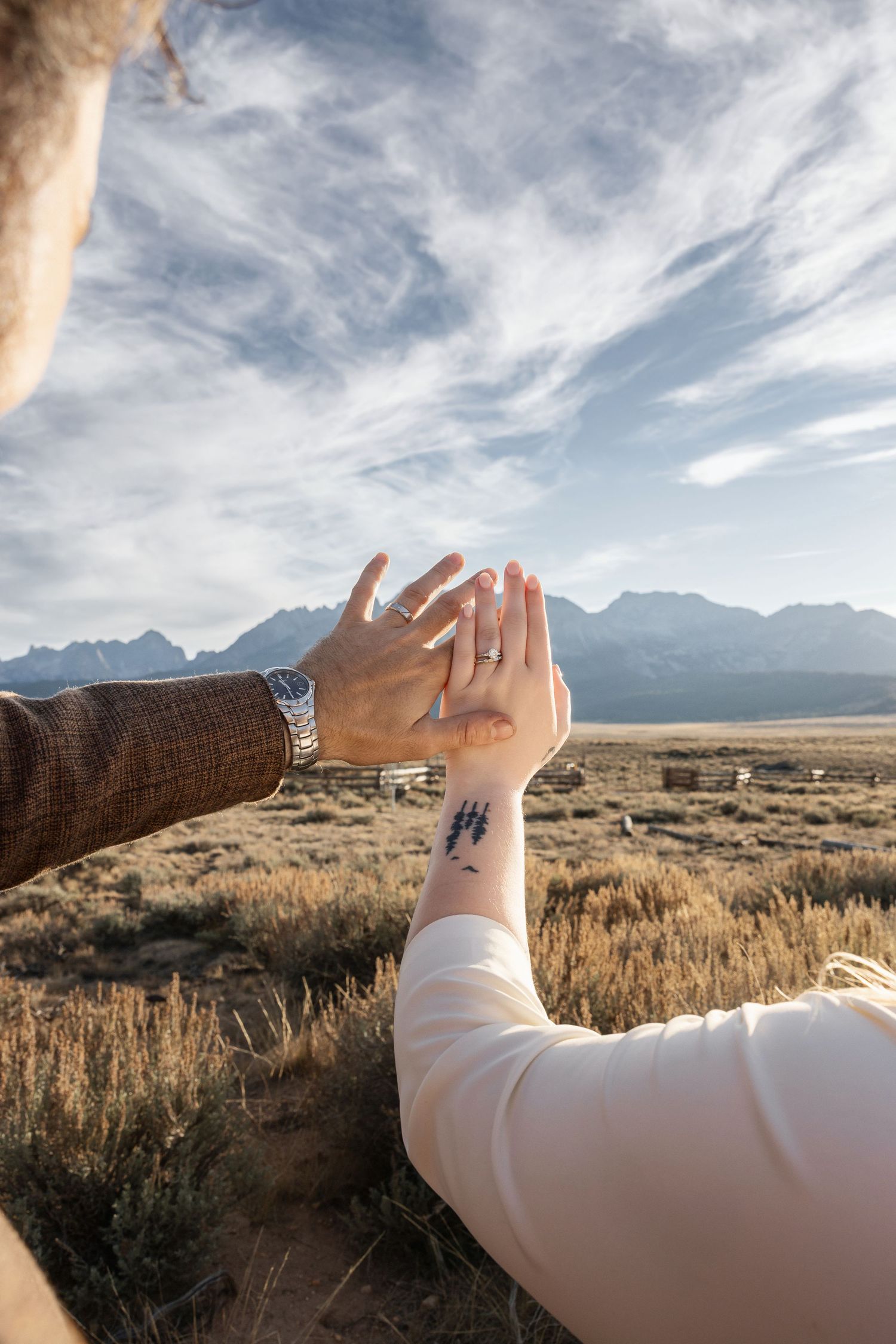 A raised arm gestures against a dramatic mountain landscape with wispy clouds at sunset.